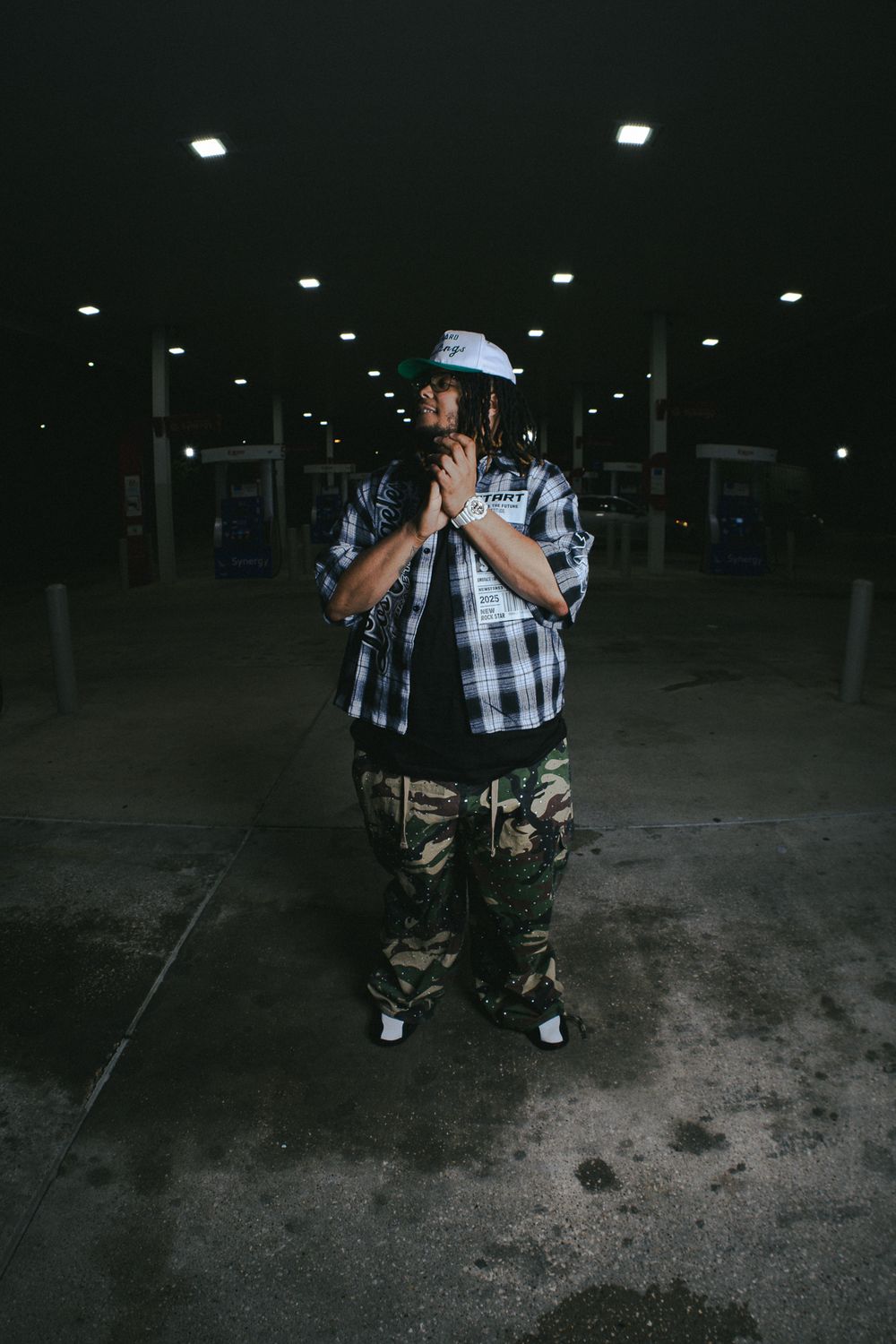 Dark parking garage scene with person in plaid shirt and camo pants posing.