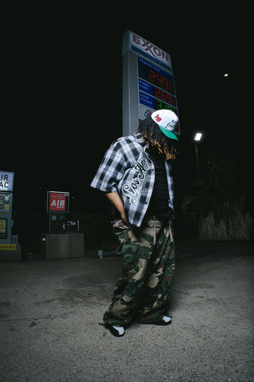 Person in plaid shirt and camo pants crouching near gas station at night.
