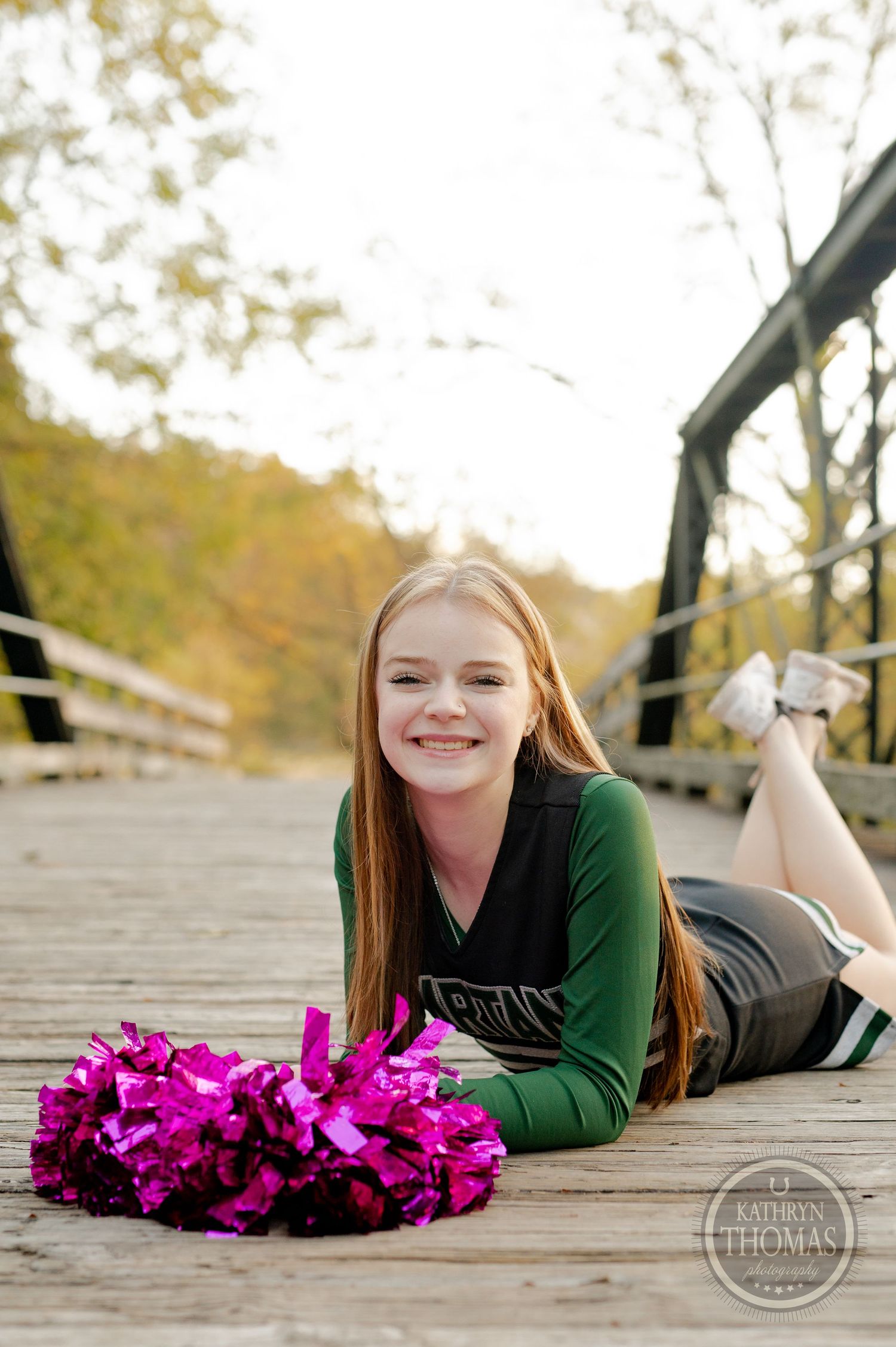 Cheerleader poses with pink pom poms lying on wooden bridge during fall photoshoot.