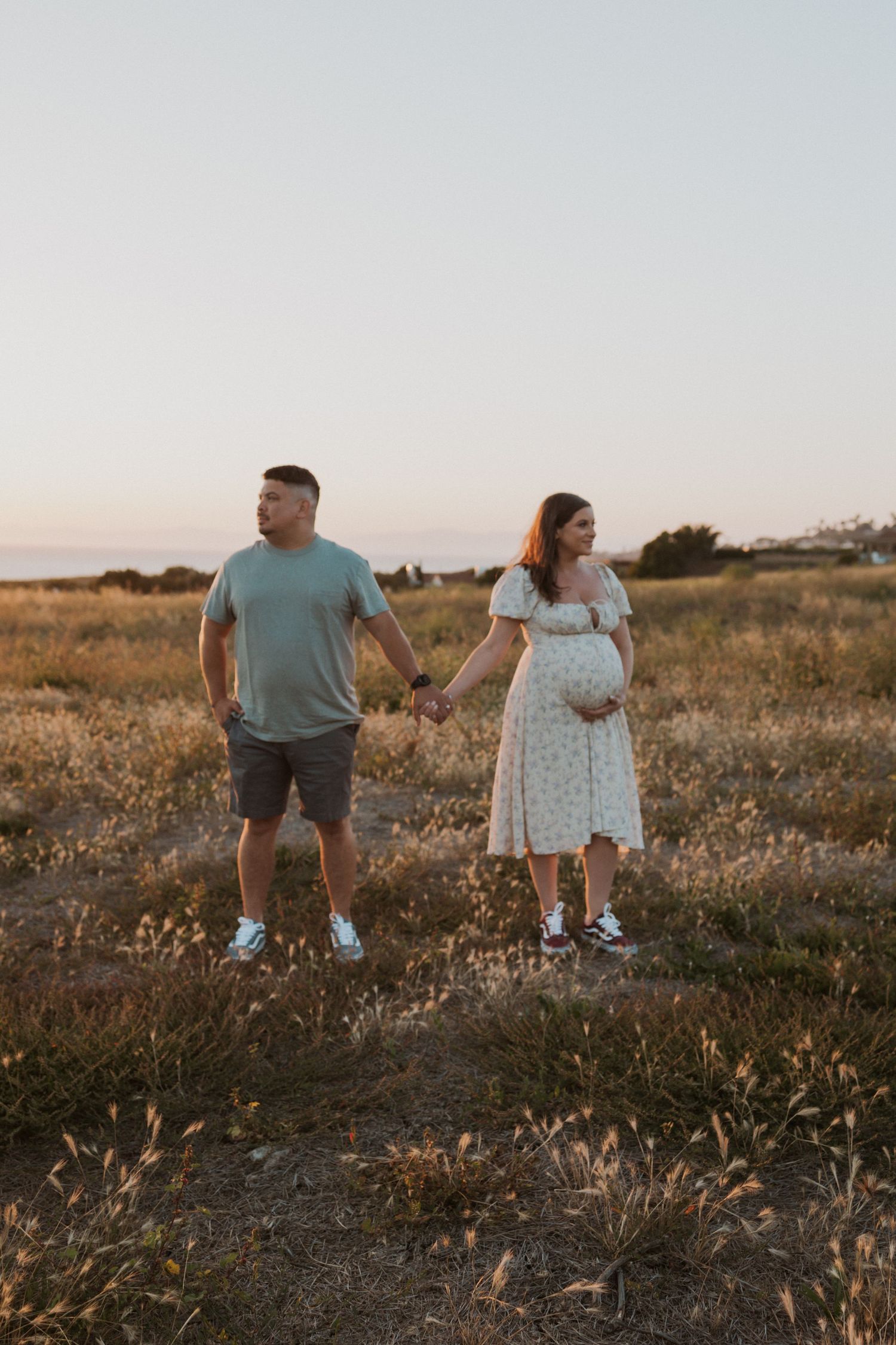 Expecting couple holds hands in golden field during sunset maternity photoshoot.