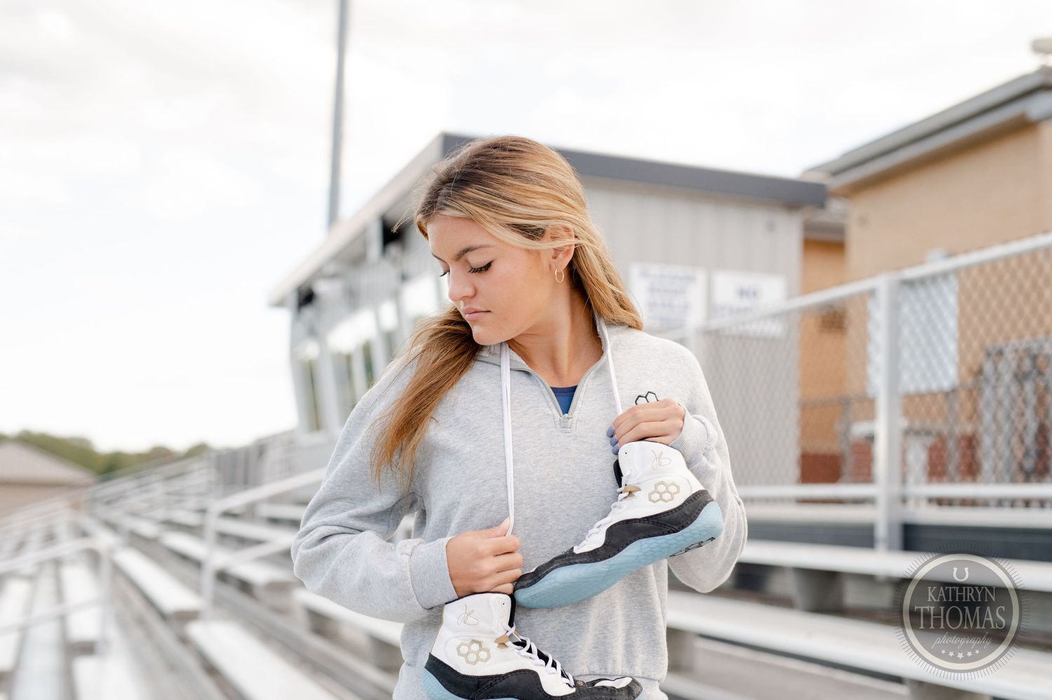 Athlete holding ice skates poses on metal bleachers wearing grey sweatshirt.