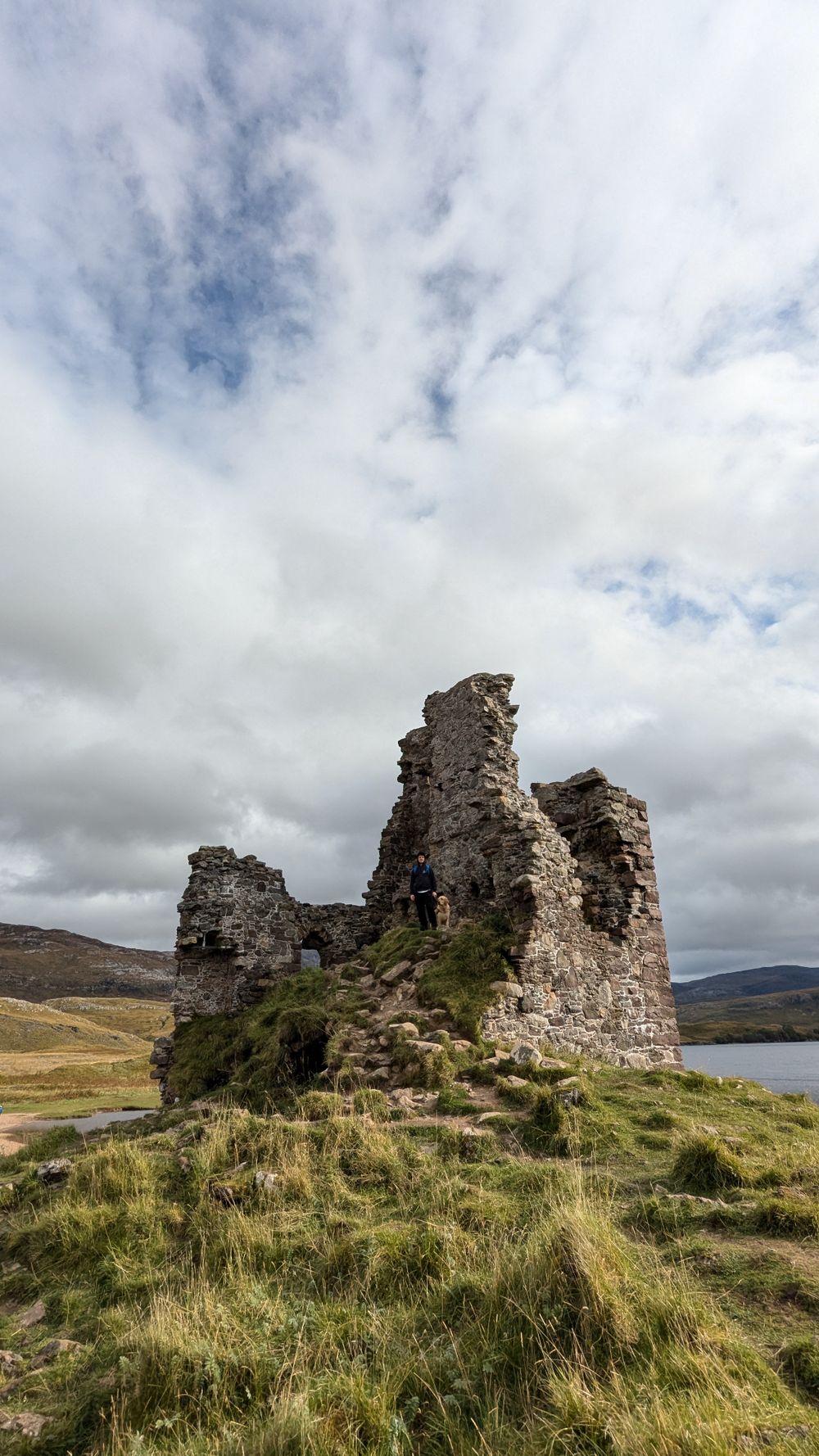 Ancient stone castle ruins stand on a grassy hill overlooking a Scottish loch under dramatic cloudy skies.