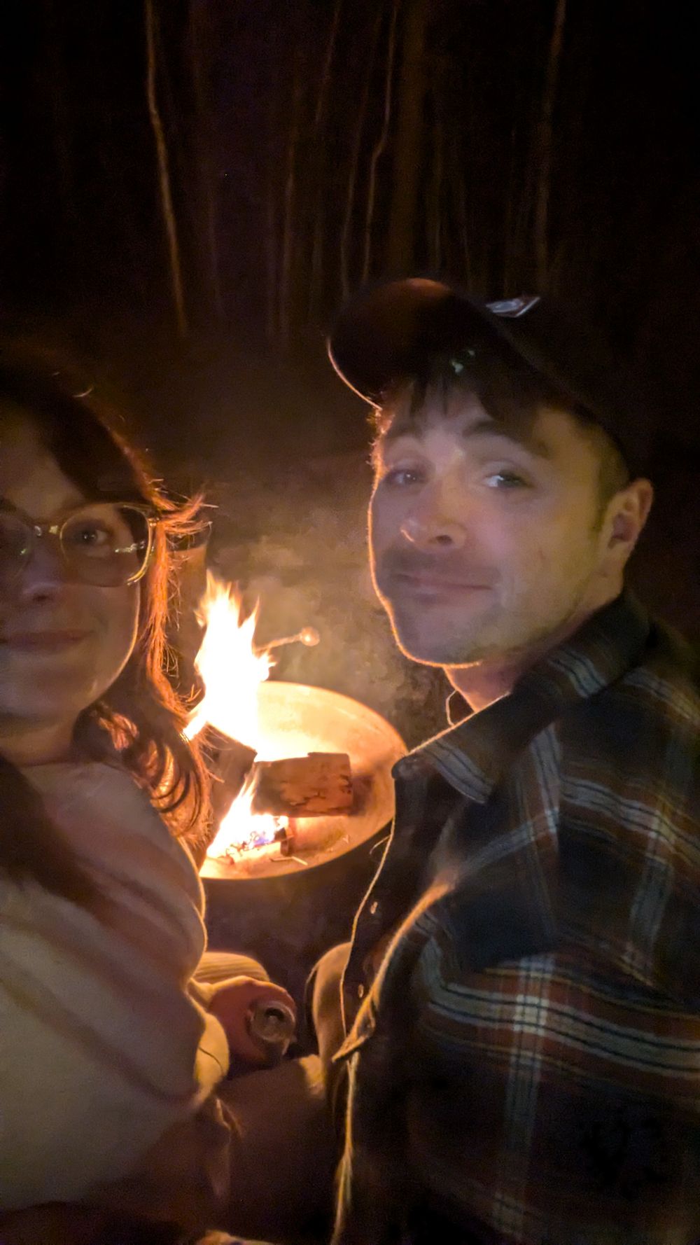 A couple sits together by a glowing campfire at night, illuminated by the warm firelight.