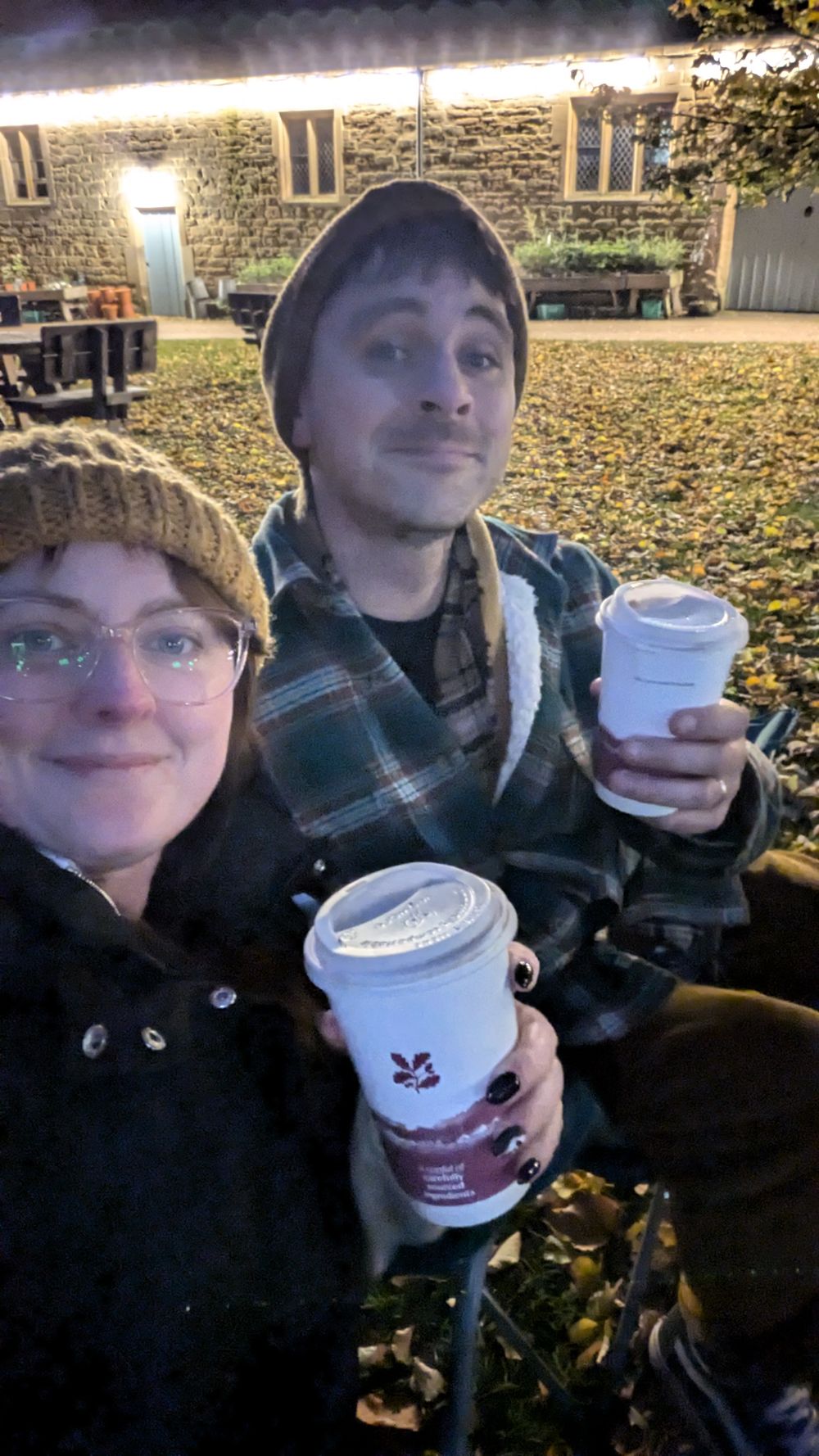 Two people enjoying hot beverages outdoors on an autumn day with fallen leaves and stone buildings in the background.