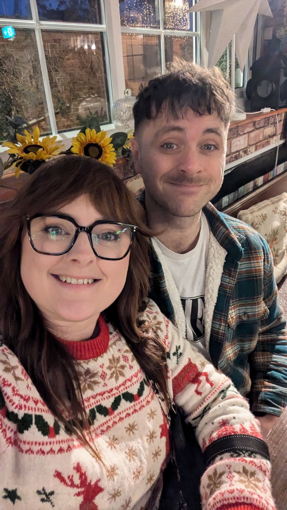 A cozy winter selfie showing two people in festive sweaters with sunflowers and brick wall decor in the background.