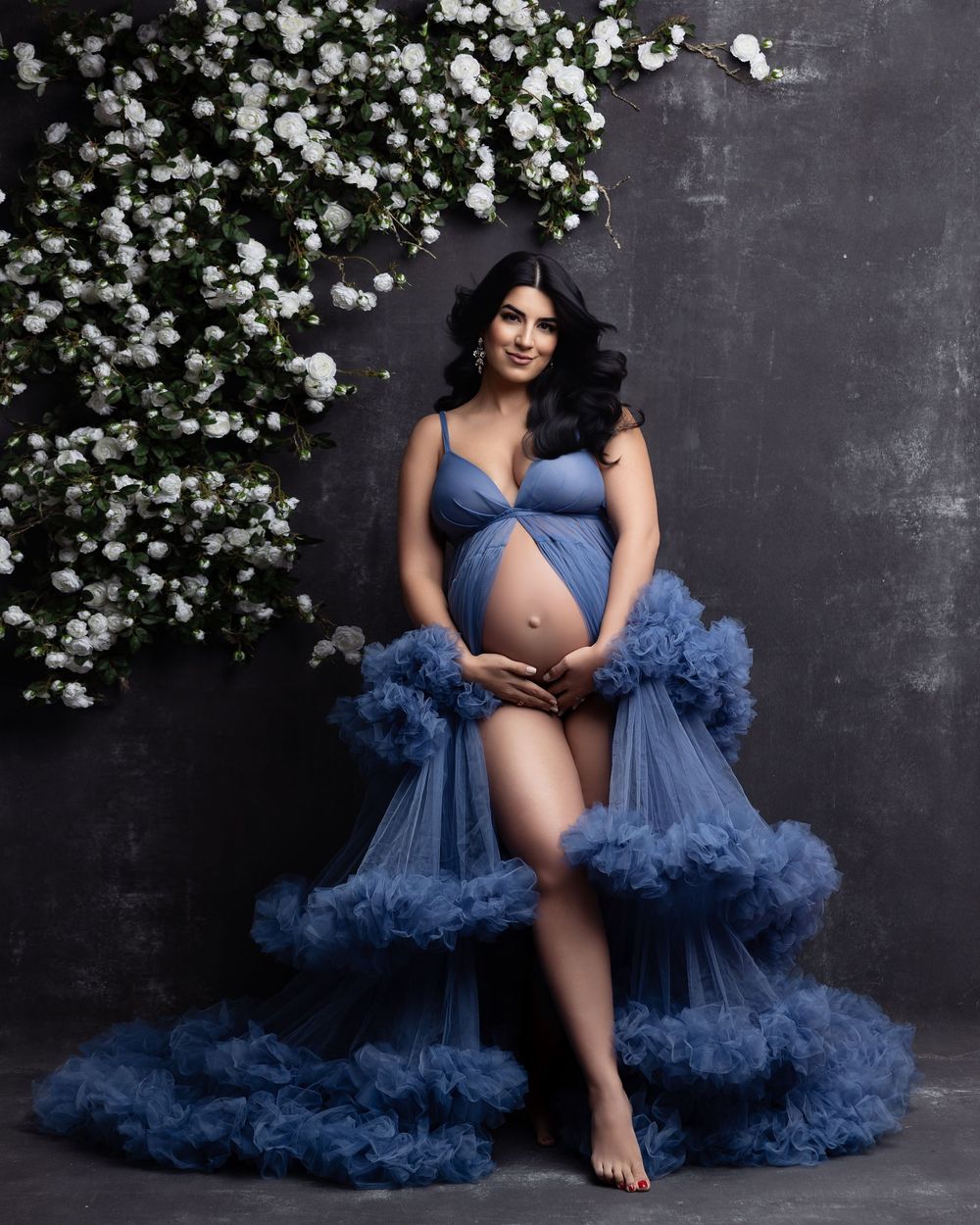 Pregnant woman in blue tulle maternity gown posing with white flowers in a studio photo shoot.