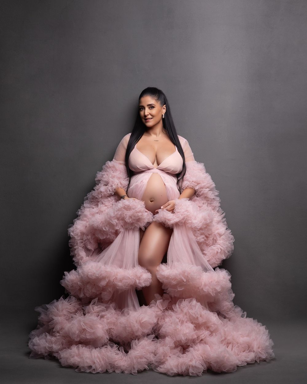 A pregnant figure poses in a flowing pink tulle robe against a grey backdrop during a maternity photoshoot.