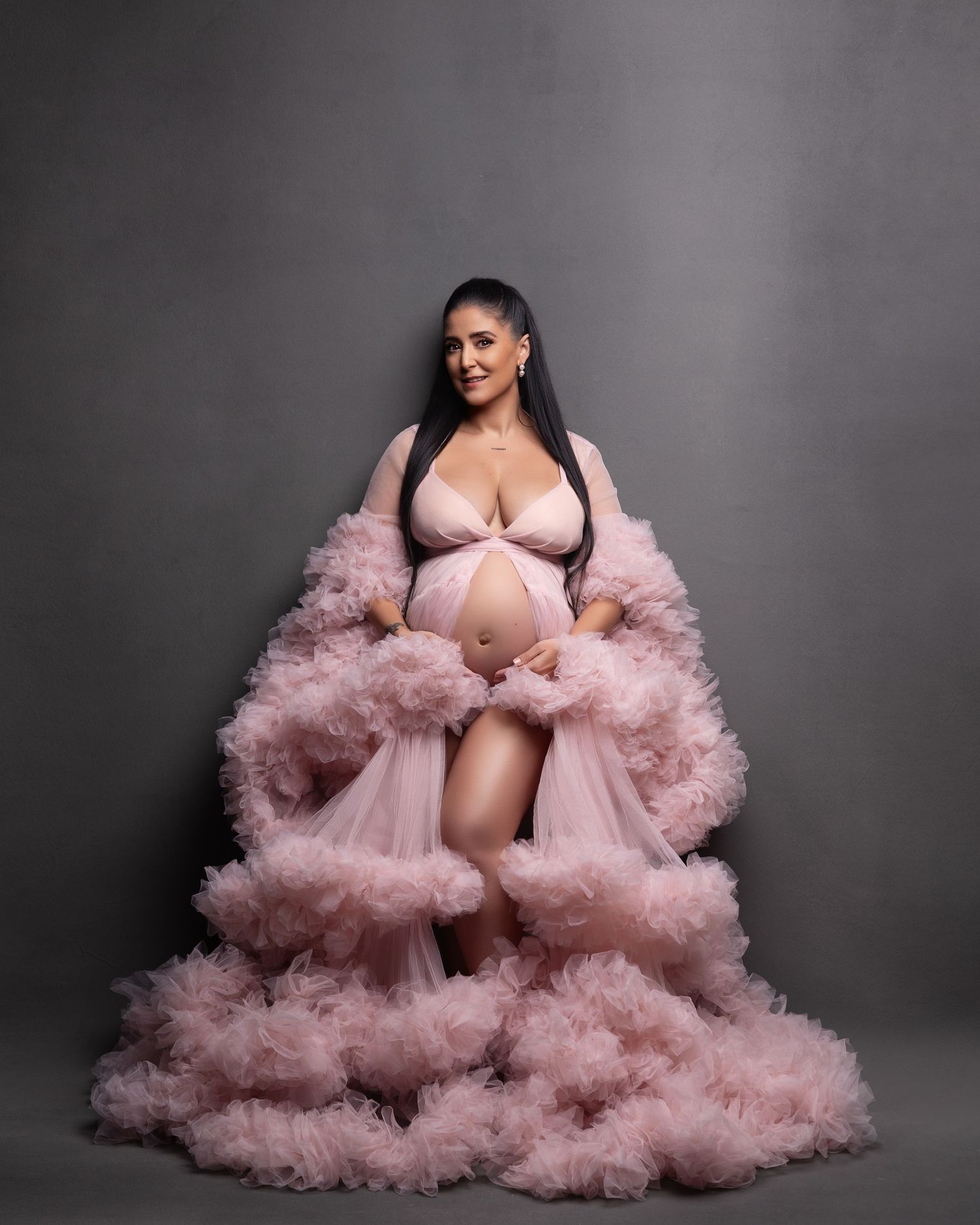 A pregnant figure poses in a flowing pink tulle robe against a grey backdrop during a maternity photoshoot.