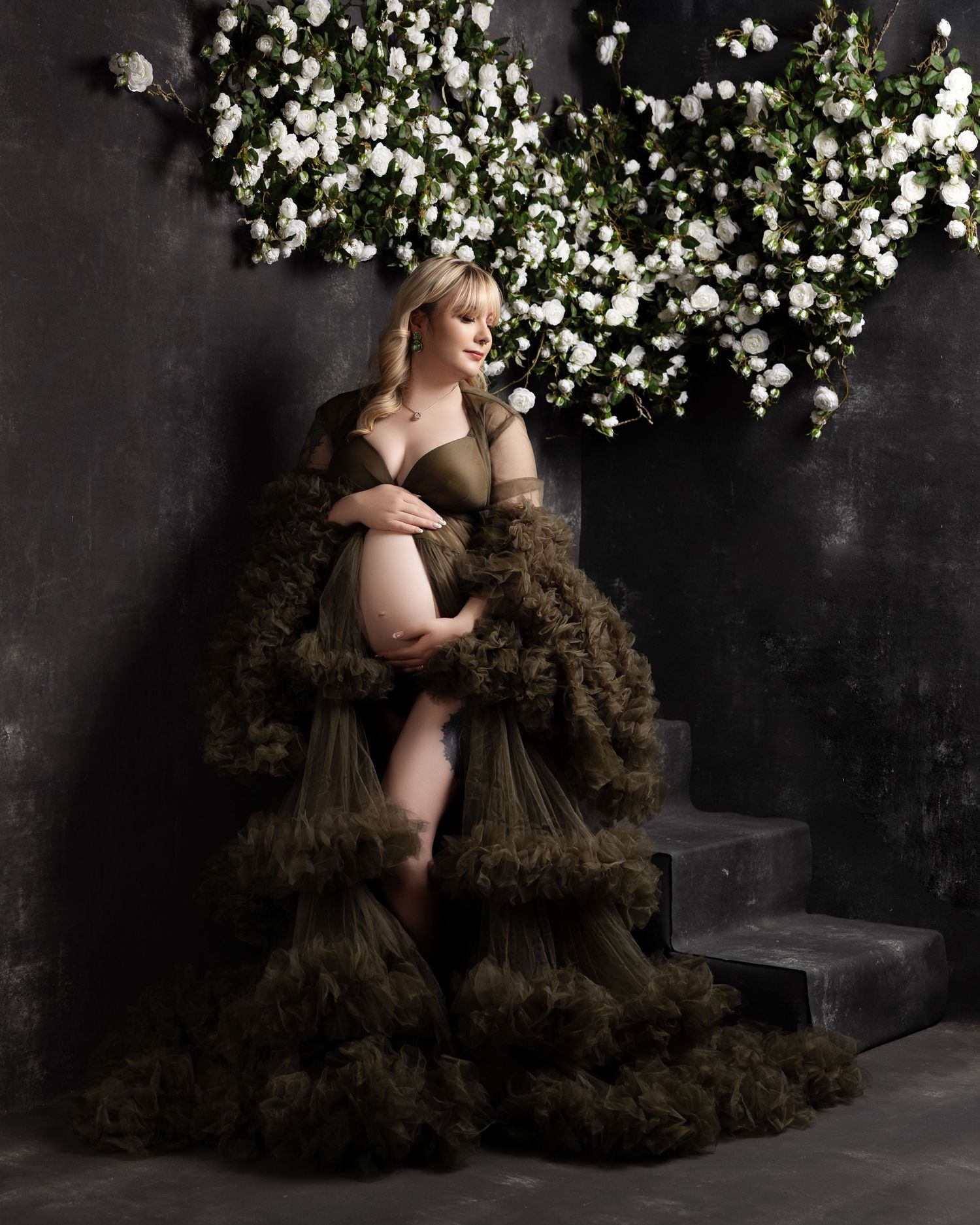 An elegant maternity portrait with flowing brown tulle fabric and white floral decorations against a dark backdrop.