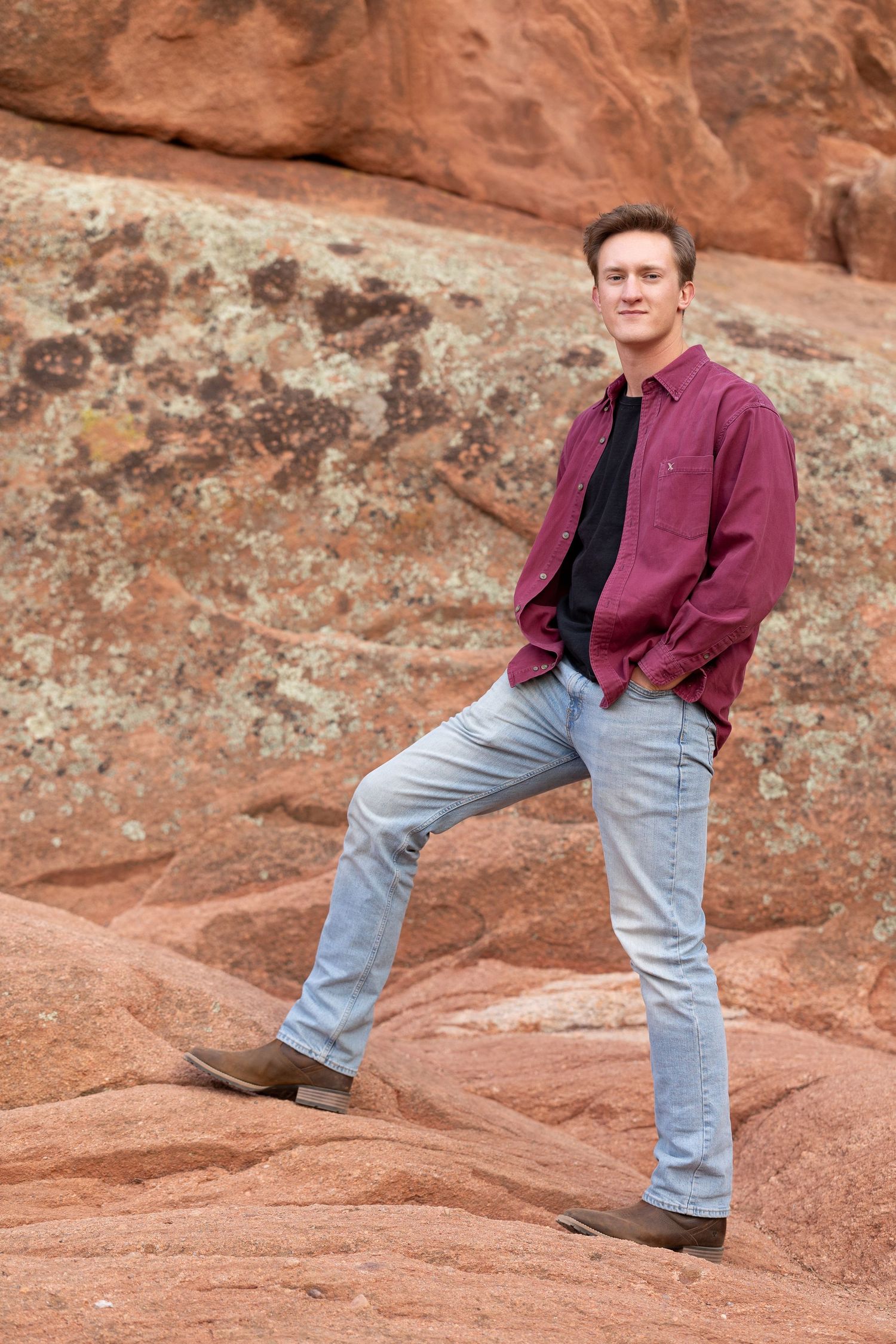 Person in burgundy shirt and jeans posing on red rock formations outdoors.