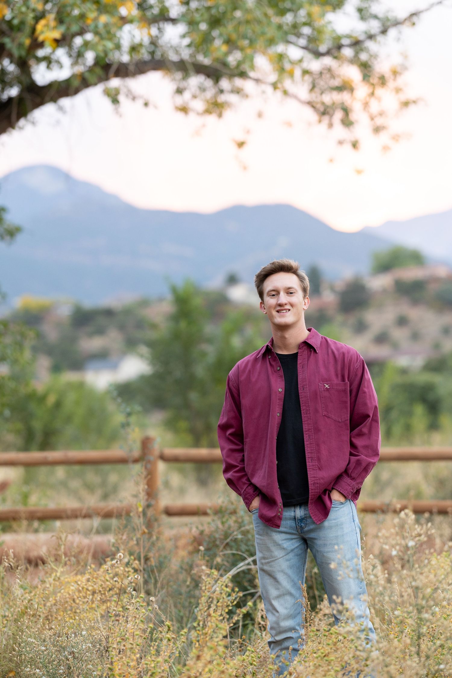 Person in burgundy shirt standing by wooden fence with mountain vista in background.