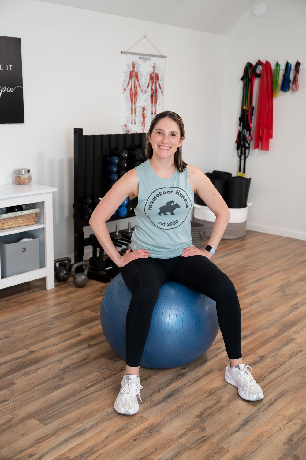 Smiling woman in Mamabear Fitness tank sitting on blue exercise ball in well-equipped home gym.