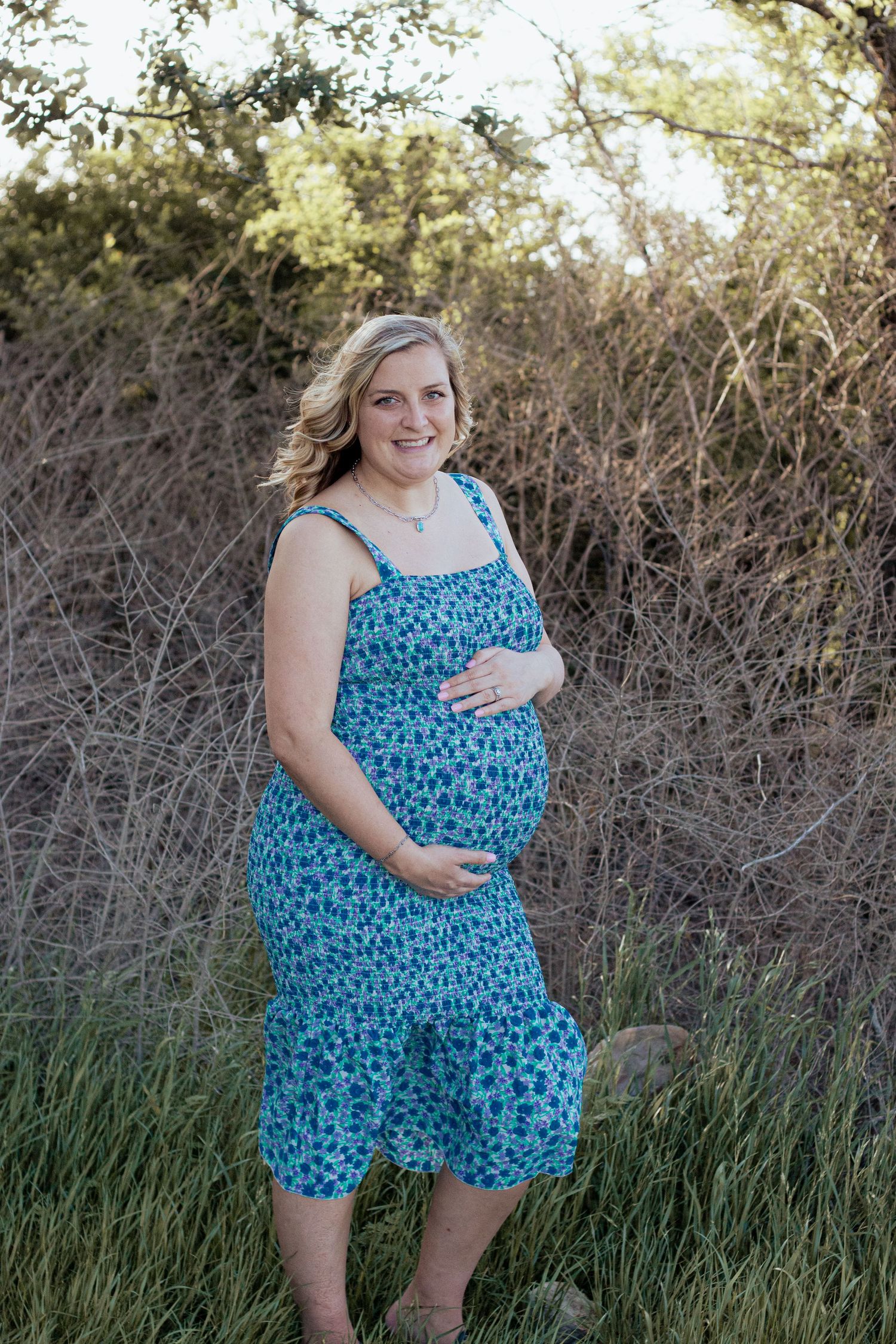 Pregnant woman in blue floral sundress stands in tall grass and natural scenery during outdoor maternity photo session.