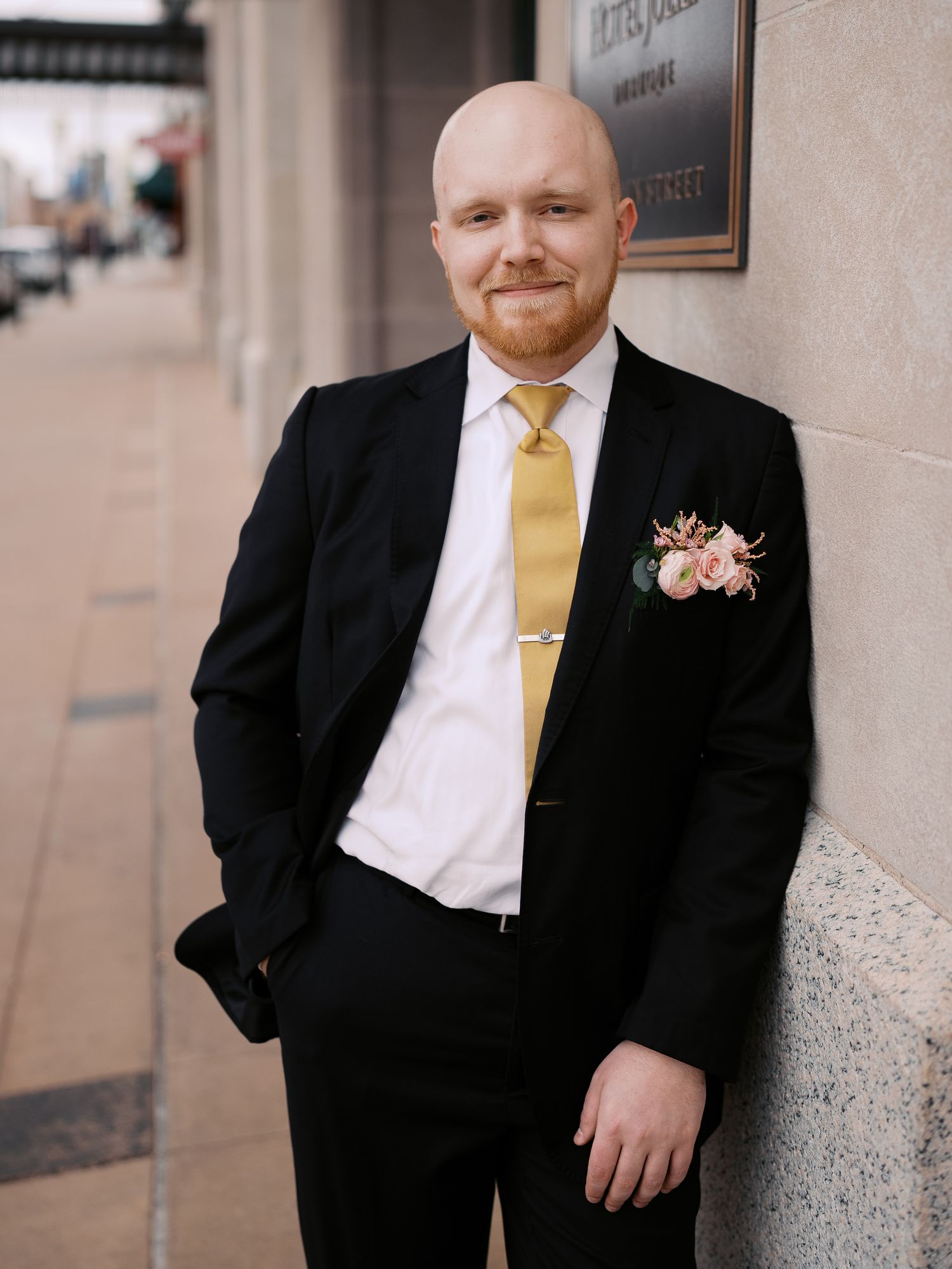 A groom in a black suit with yellow tie and floral boutonniere poses against a wall during a wedding.