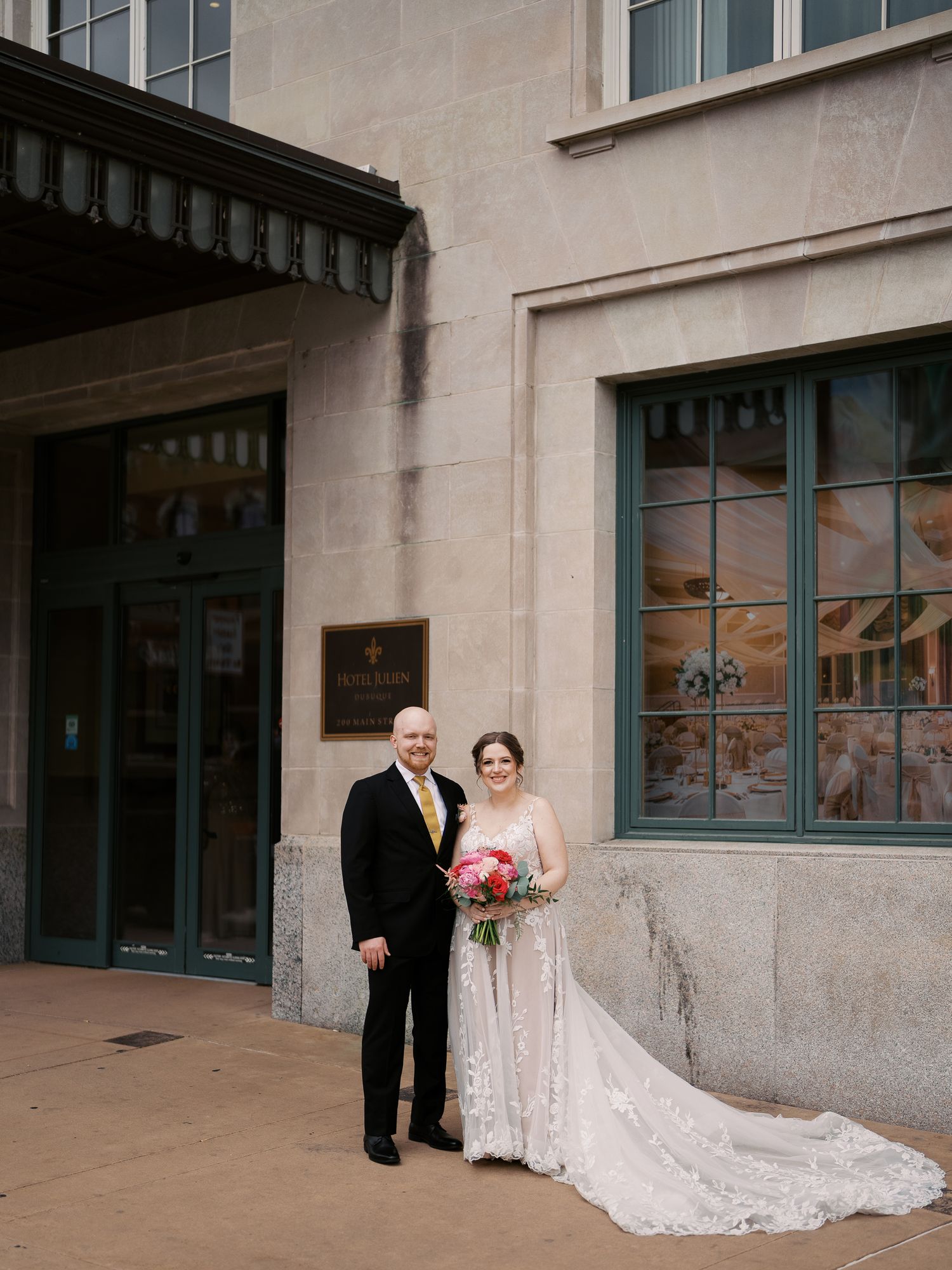 A couple in wedding attire stands outside an elegant building with glass windows and a stone facade.