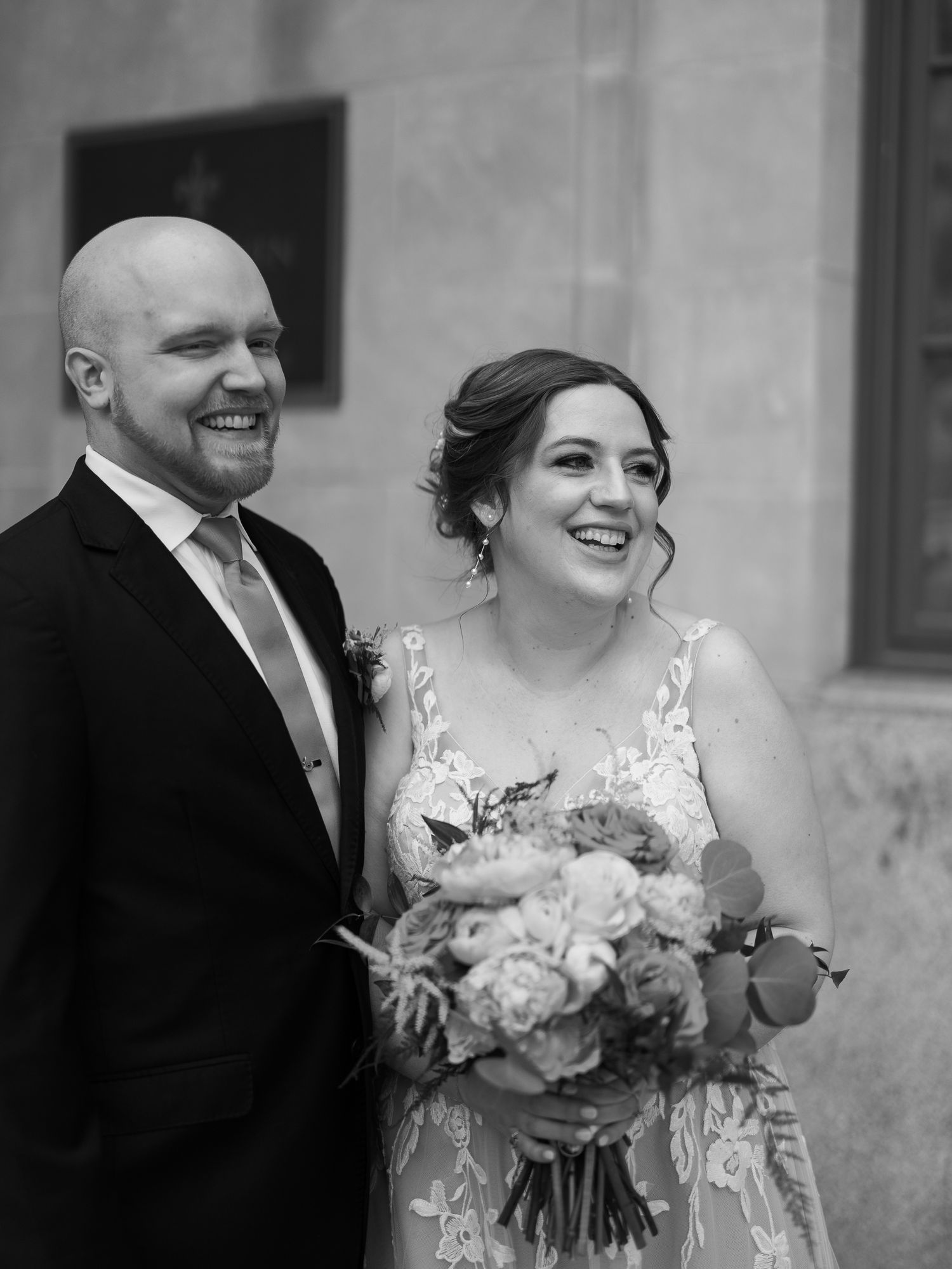 A newlywed couple shares a joyful moment in black and white as they pose with a bouquet of pink and white flowers.