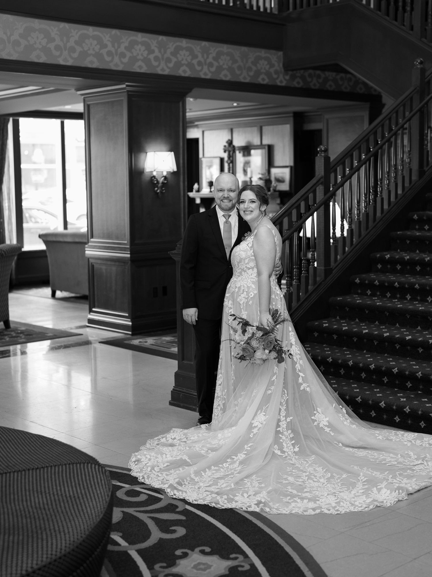 Elegant black and white photograph of a couple posing by a grand staircase in formal wedding attire.