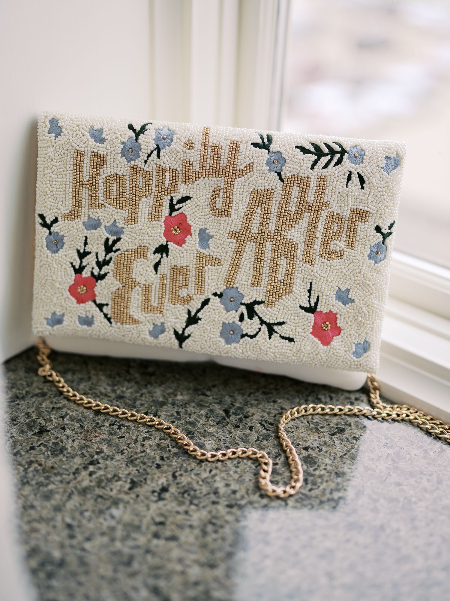 White beaded clutch purse with floral embroidery and 'Happy Ever After' text features a delicate gold chain strap.