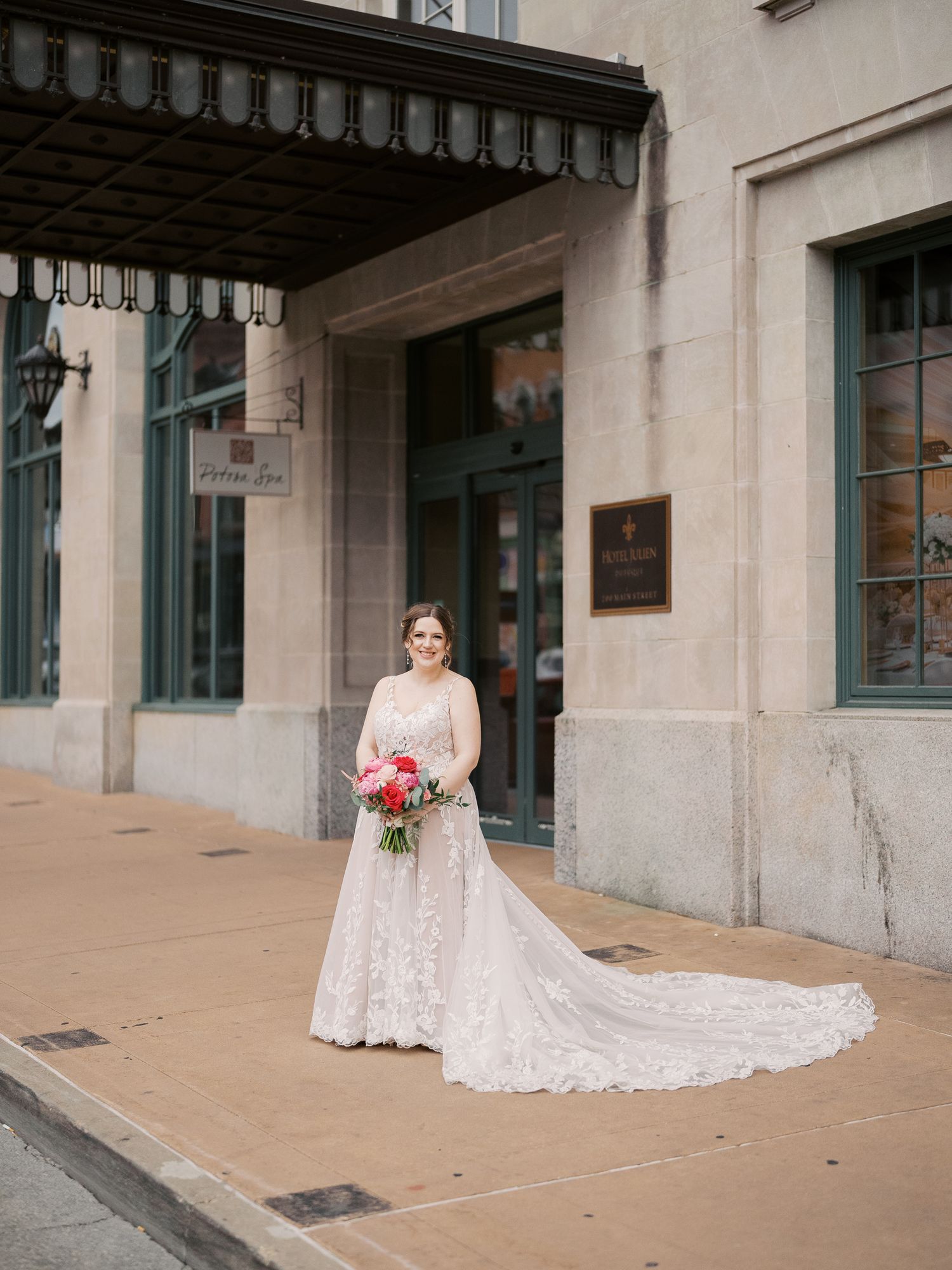 A person in a lace wedding dress with a long train stands in front of a historic building entrance holding pink flowers.