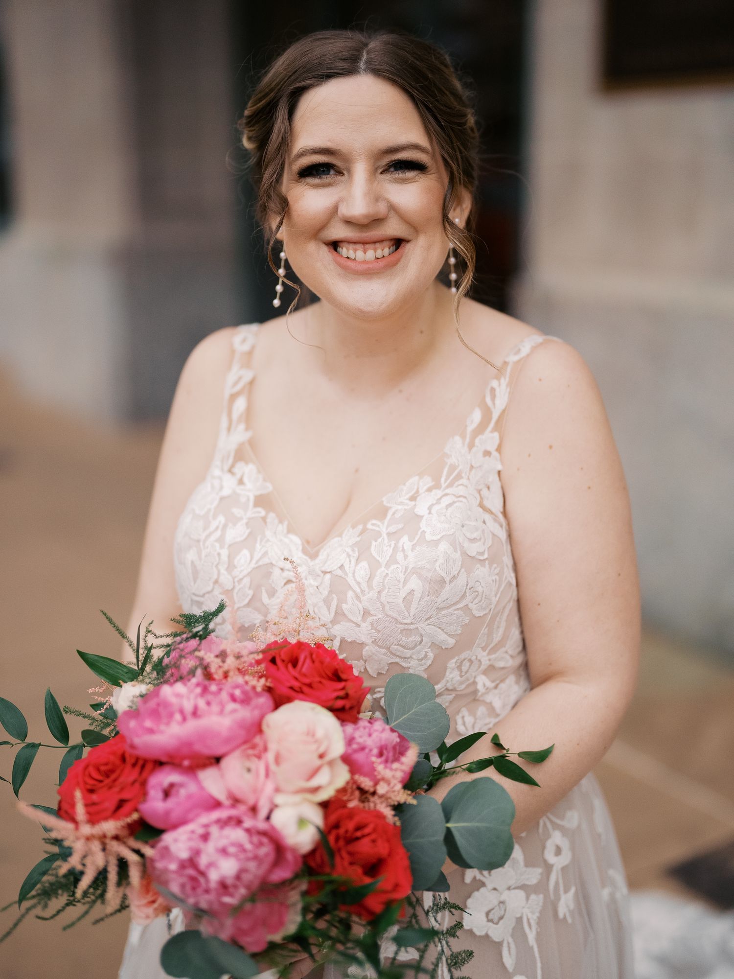 A bride in a white lace gown holds a vibrant bouquet of pink and red flowers with eucalyptus greenery.