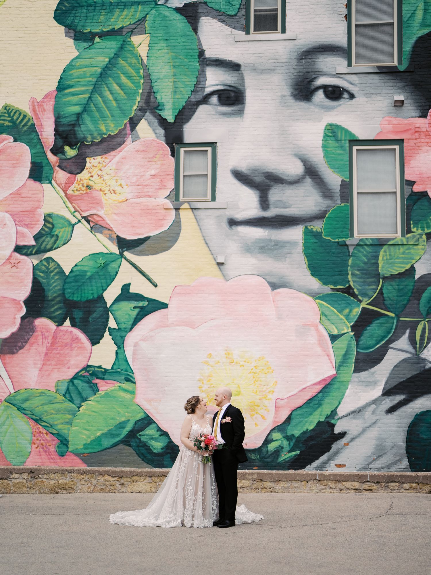 A newlywed couple poses in front of a large floral mural with pink blooms and green leaves painted on a white wall.