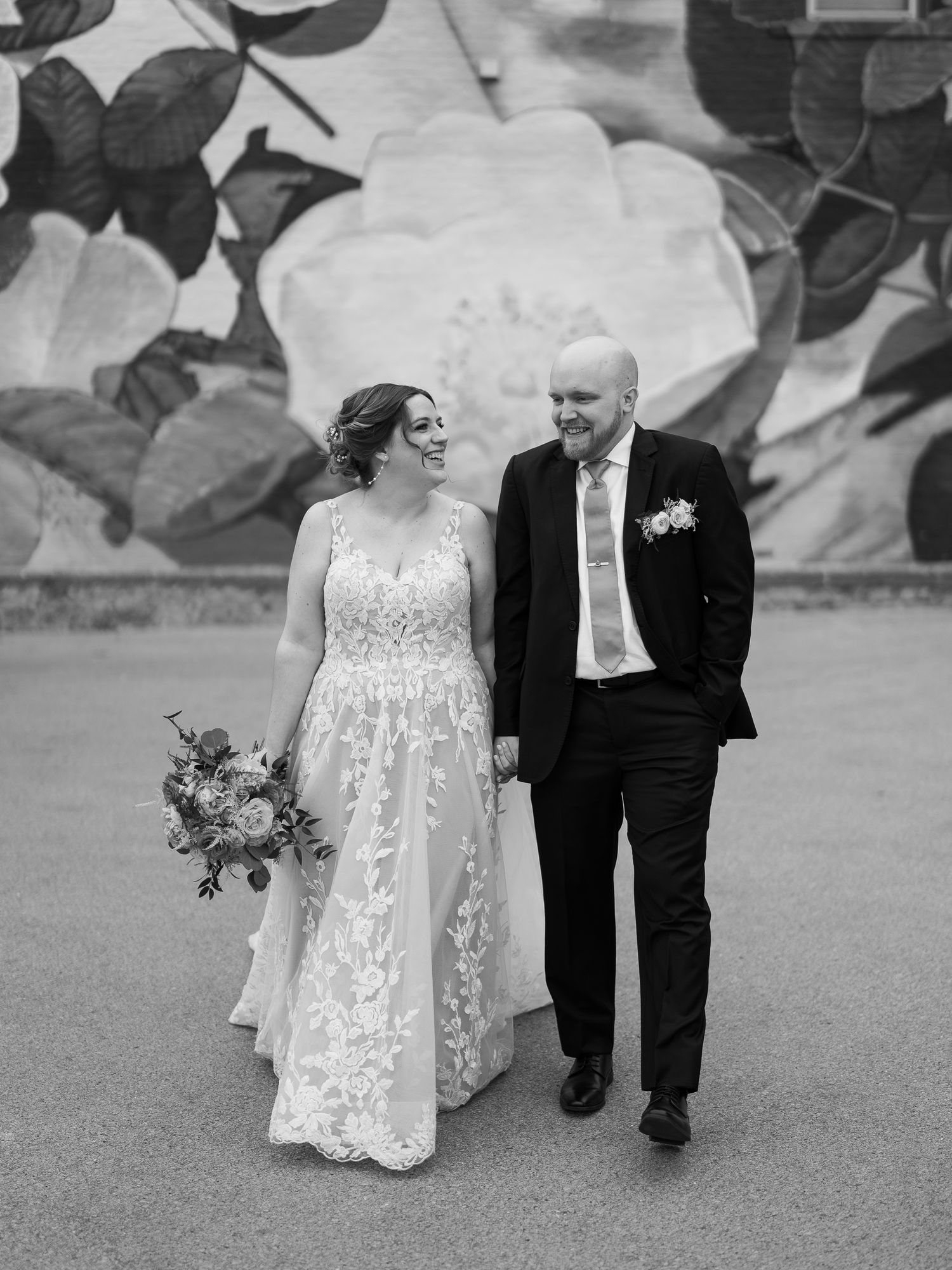 Black and white wedding photo of couple walking together, bride in lace gown and groom in dark suit with boutonniere.