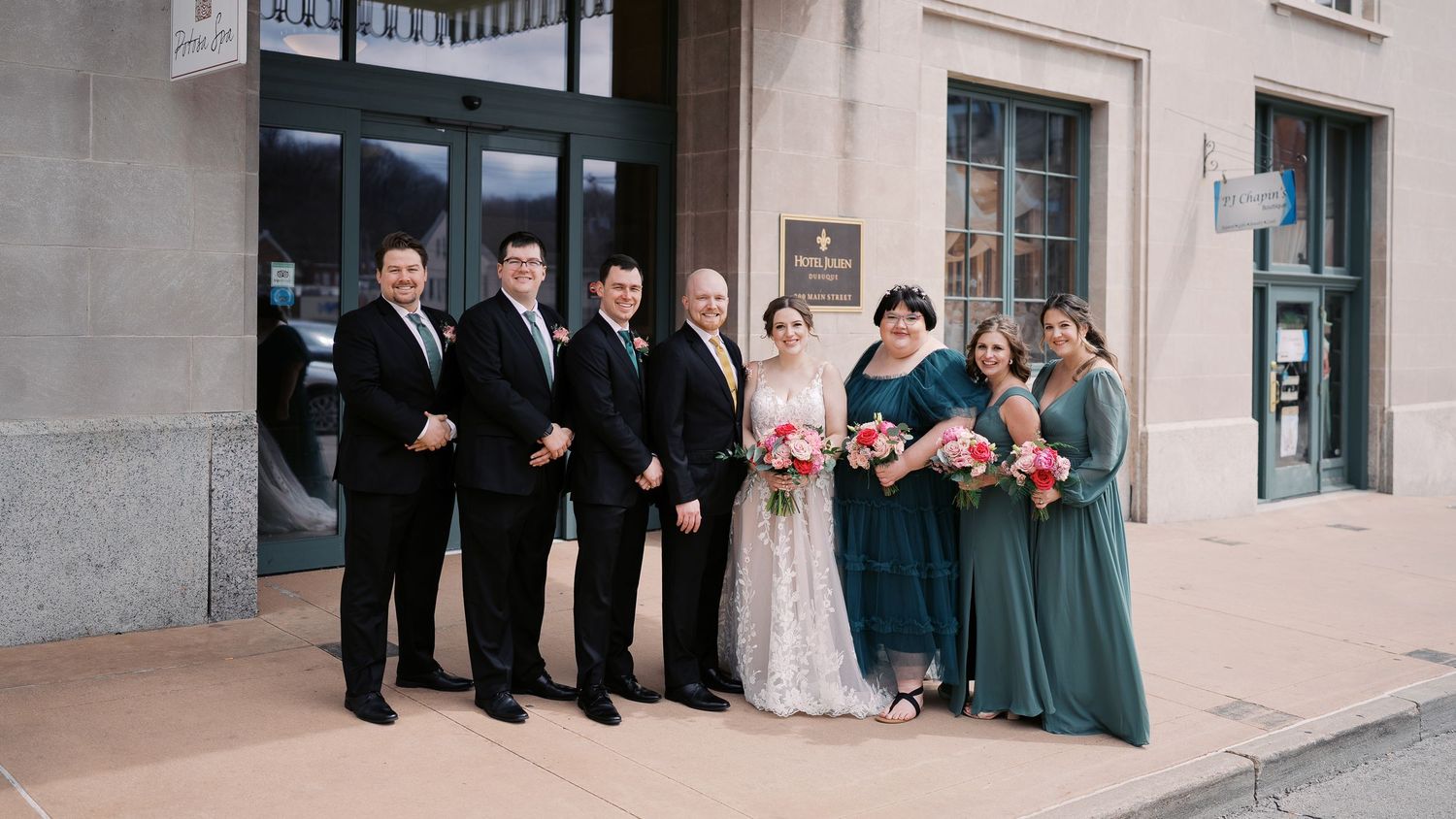 A wedding party poses outside a historic building with people in formal black suits and teal dresses.