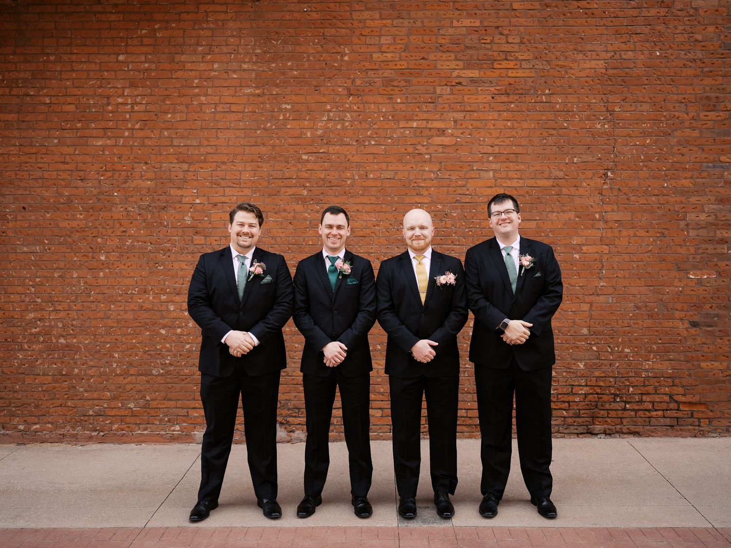 Four groomsmen wearing matching black suits and ties stand in a line against a rustic red brick wall.