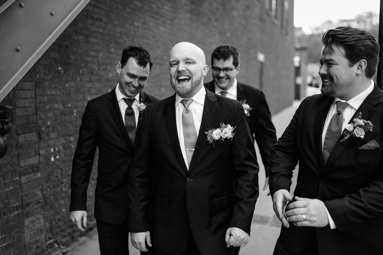 A joyful candid black and white moment with groomsmen laughing together against a brick wall during a wedding.