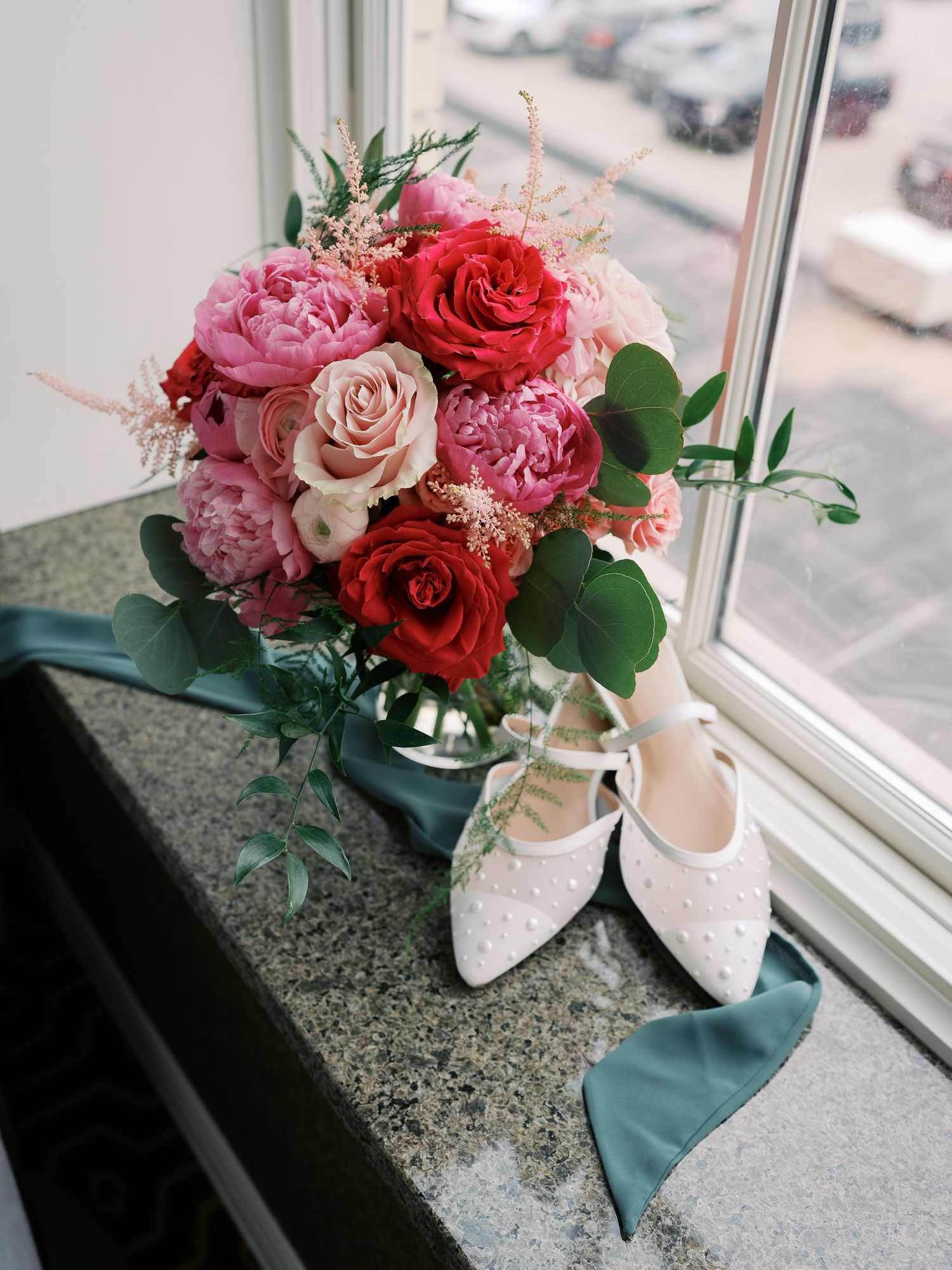 A romantic bouquet of red and pink roses paired with delicate white lace bridal shoes sits by a window.