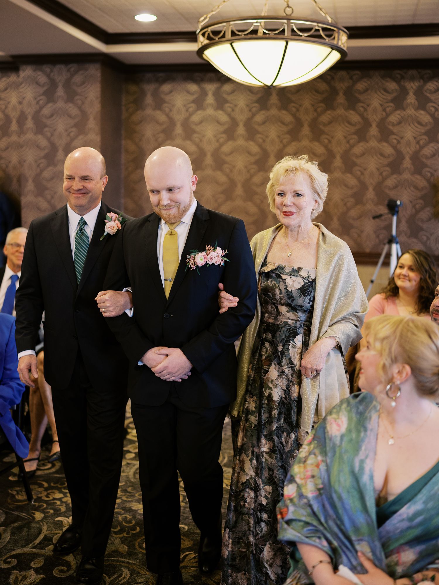 Three people walk arm in arm in formal attire at an indoor event with patterned wallpaper and elegant lighting.