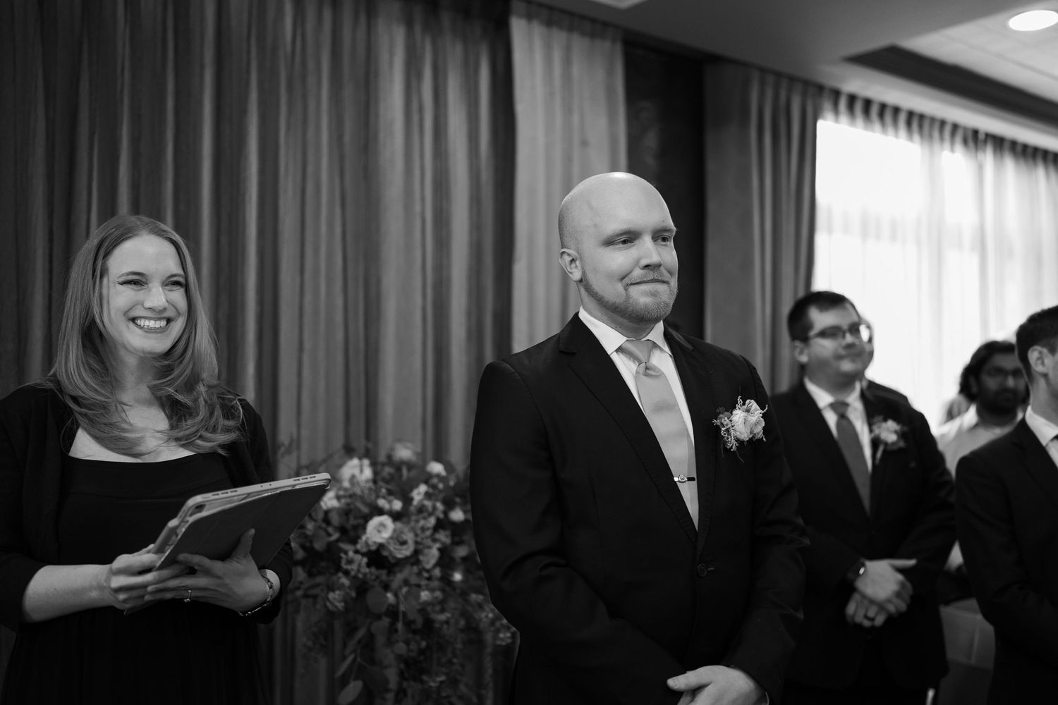 Black and white photo showing people gathered at a formal event with dramatic curtains in the background.