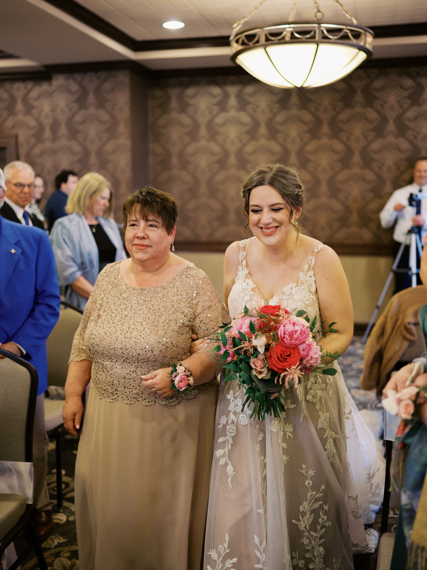 Two people walk down the aisle at an indoor wedding ceremony with elegant wallpaper and lighting decor.