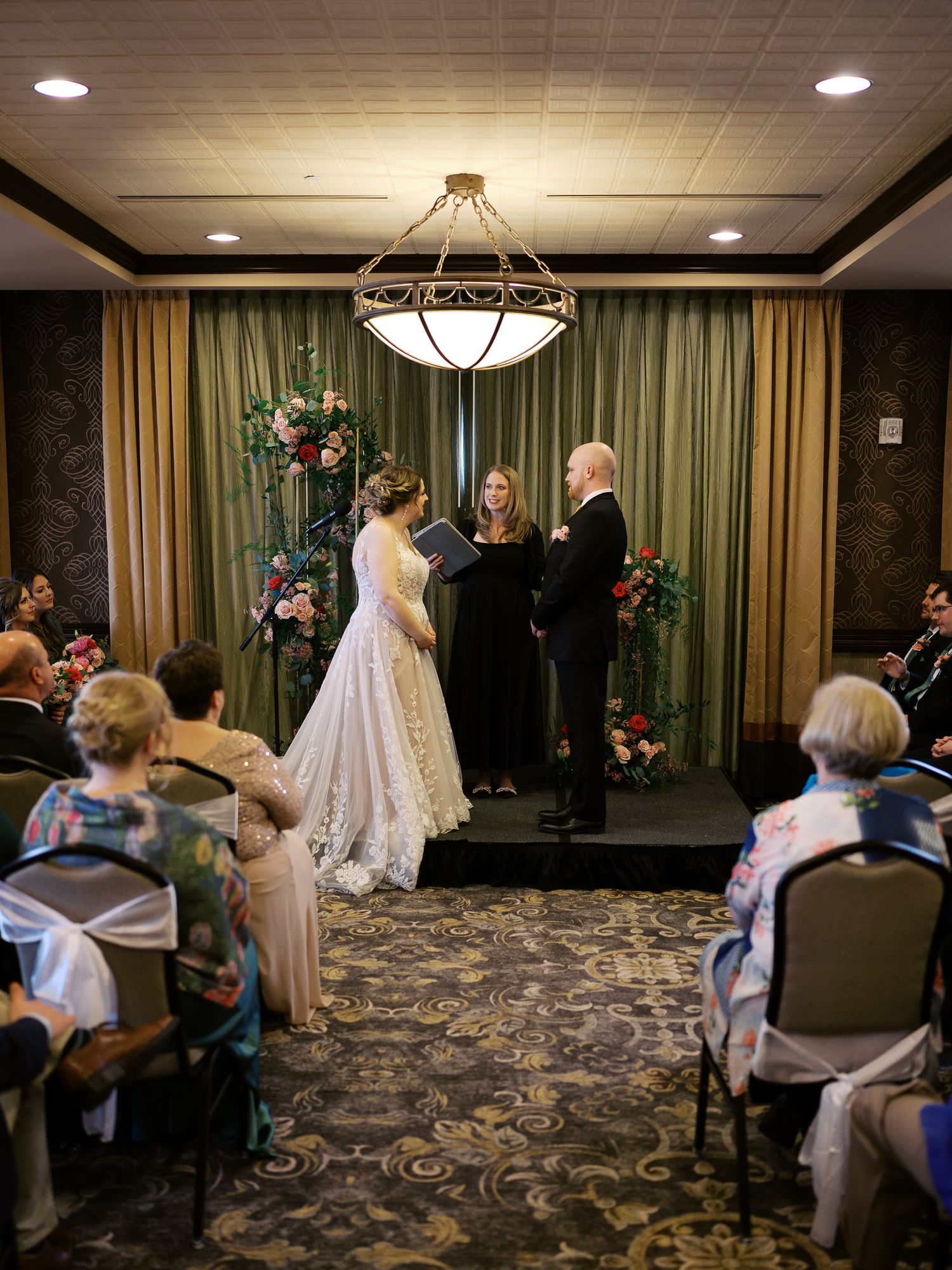 Wedding ceremony in an elegant indoor venue with floral arrangements and a crystal chandelier overhead.