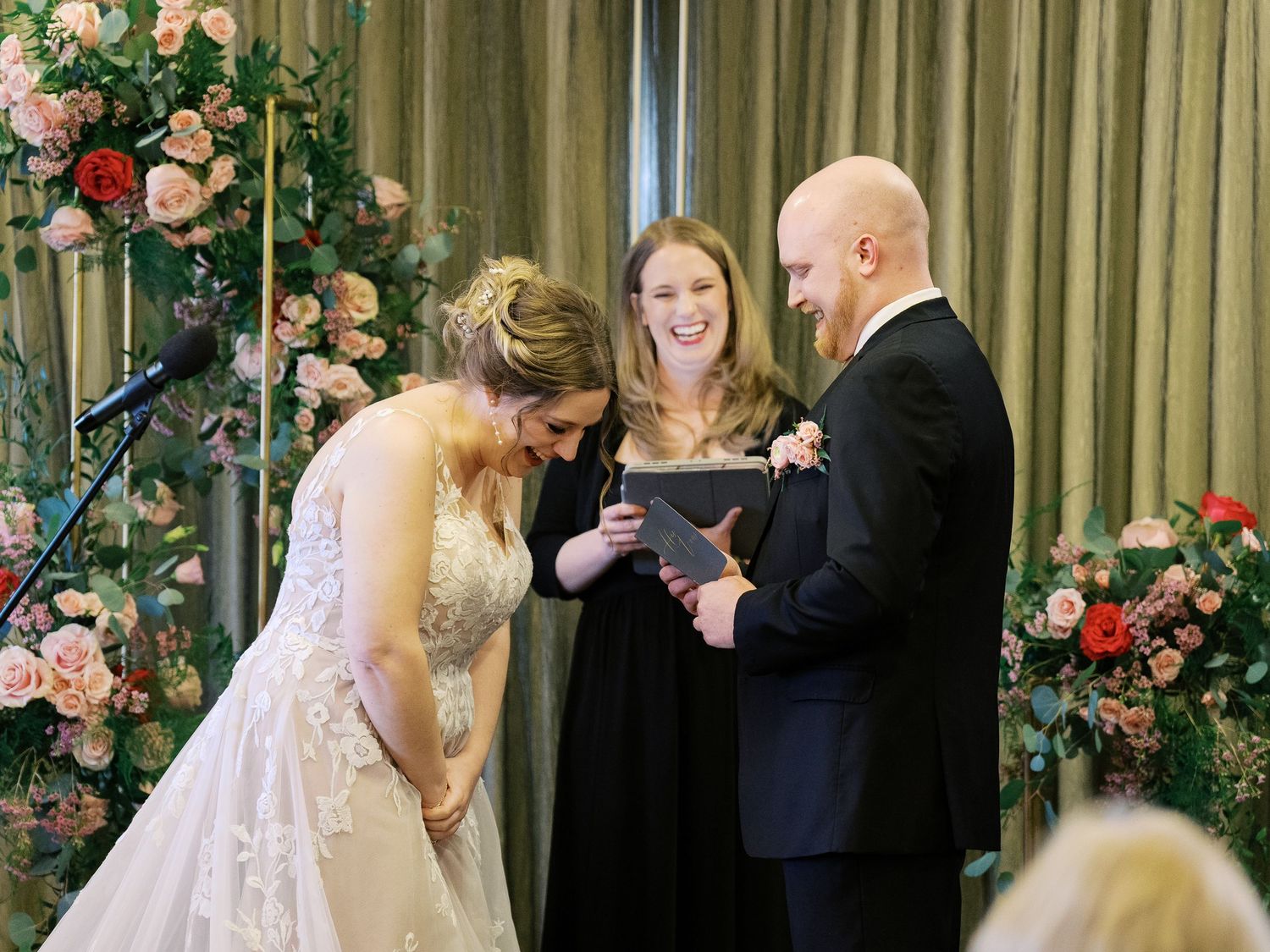 Indoor wedding ceremony with floral arch decorations in pink and white roses against green curtains.