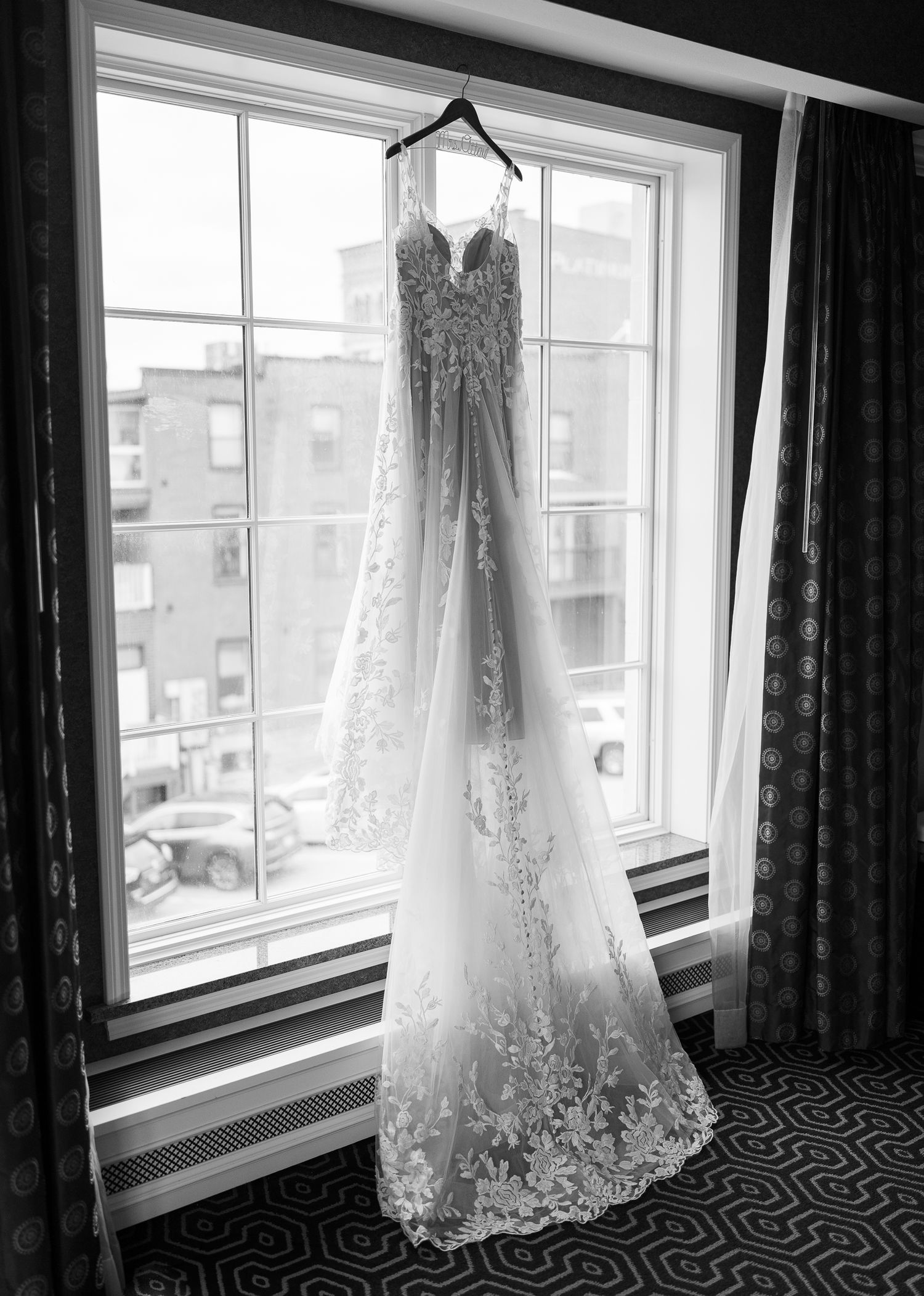 A delicate lace wedding dress hangs in silhouette against a bright window with a patterned carpet and city view beyond.