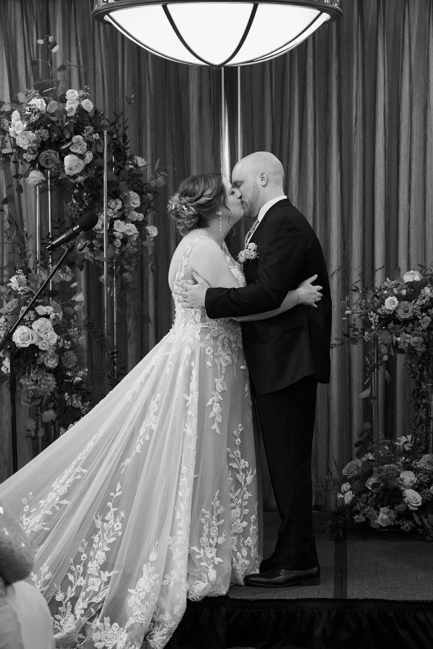 Bride in lace gown and groom share an intimate moment under dramatic lighting surrounded by floral arrangements.