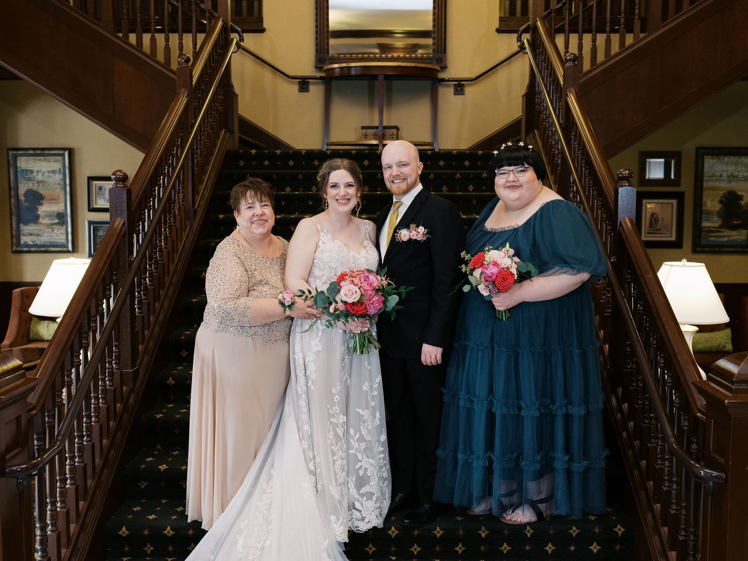 Wedding party in formal attire pose on elegant staircase with dark wood railings and patterned carpet.