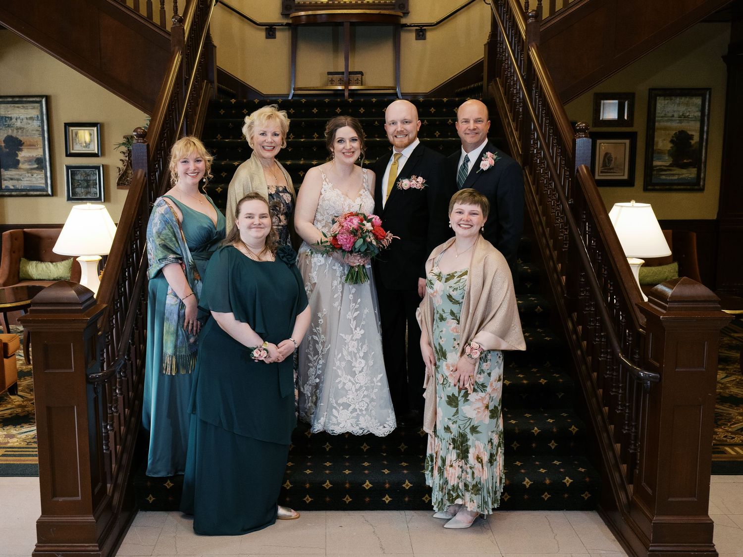 A wedding party poses on an elegant wooden staircase in a vintage-style interior with ornate decor and warm lighting.
