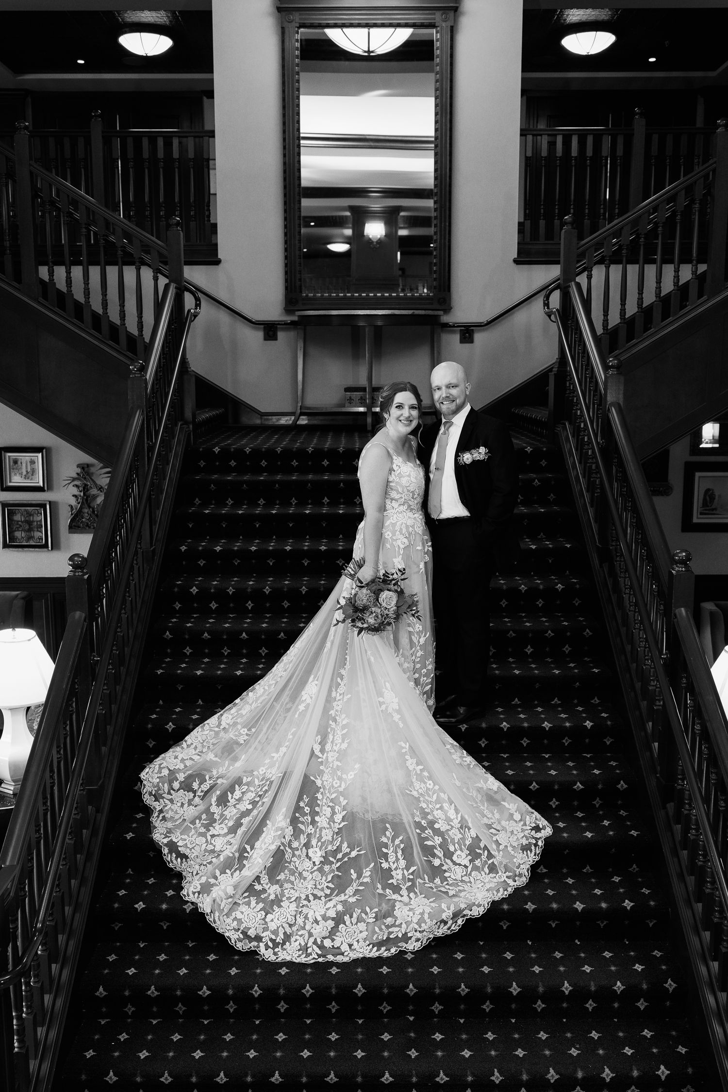 A wedding couple poses on an elegant dark carpeted grand staircase with dramatic black and white lighting.