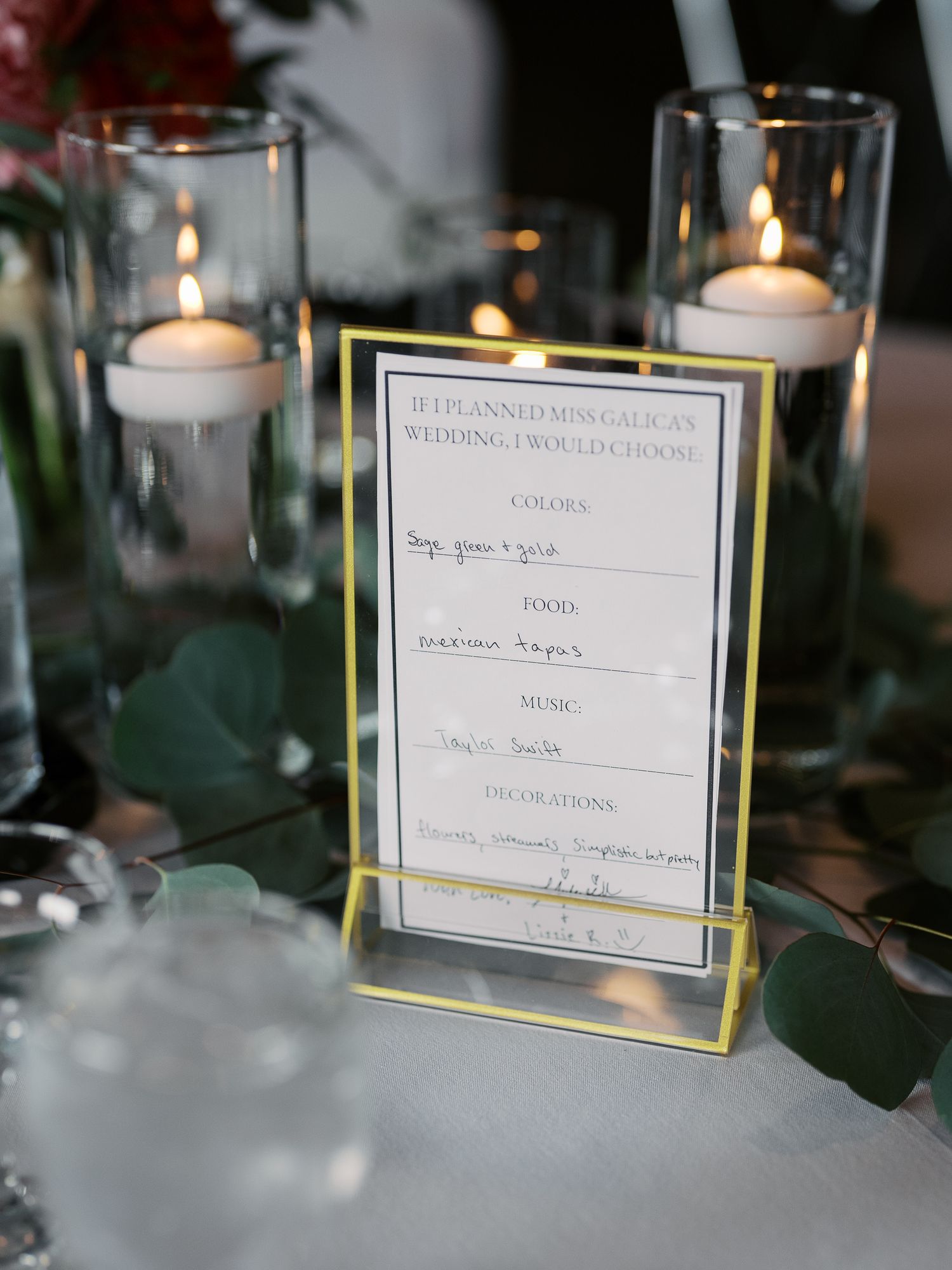 An elegant wedding menu card in a gold frame displayed on a table with glowing white votive candles and greenery.