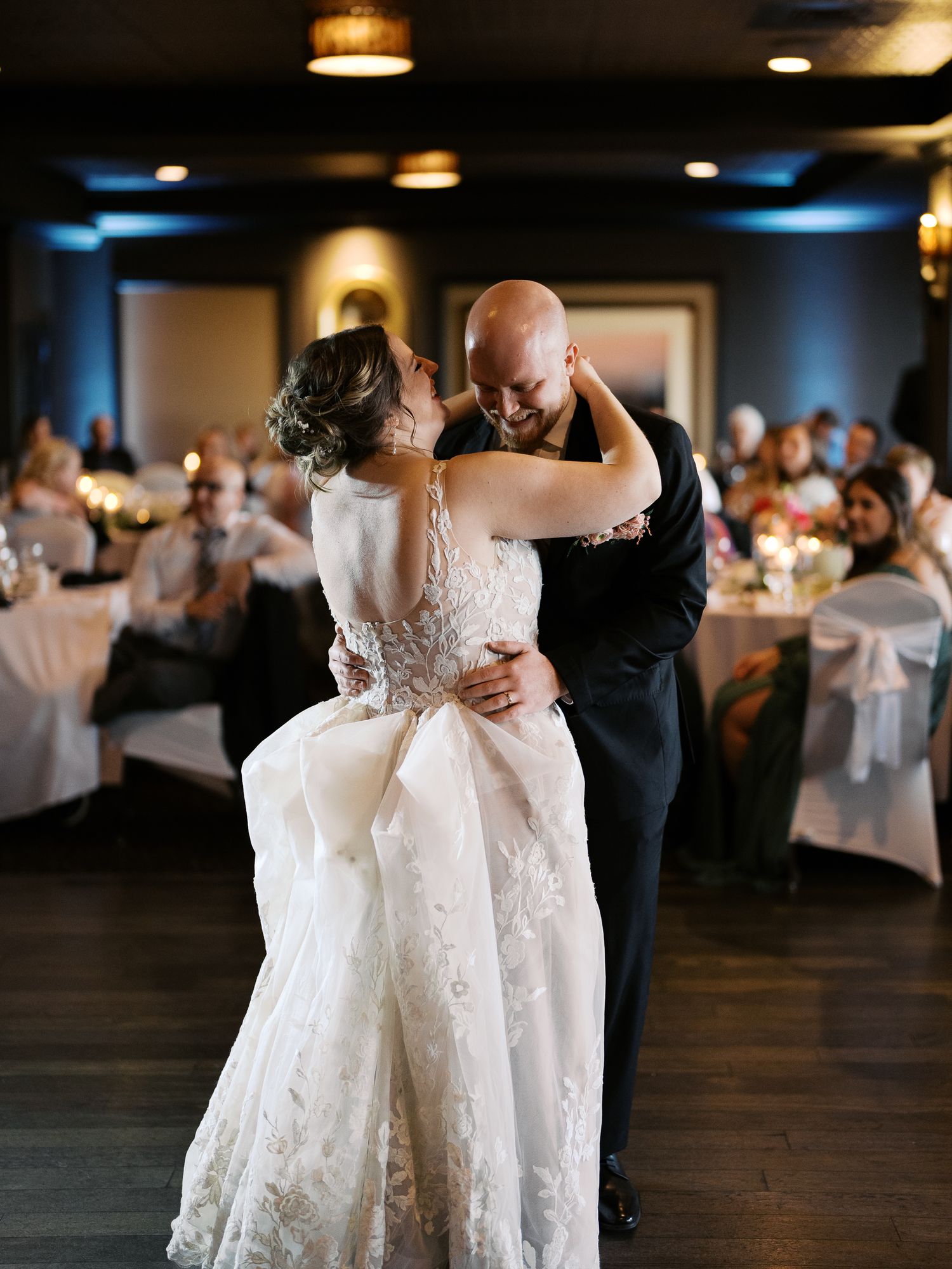 A bride and groom share their first dance at a wedding reception in an elegant ballroom with white table decor.