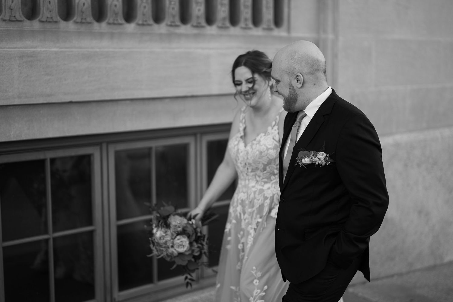 A joyful couple in formal wedding attire smile while walking together outside an elegant building.