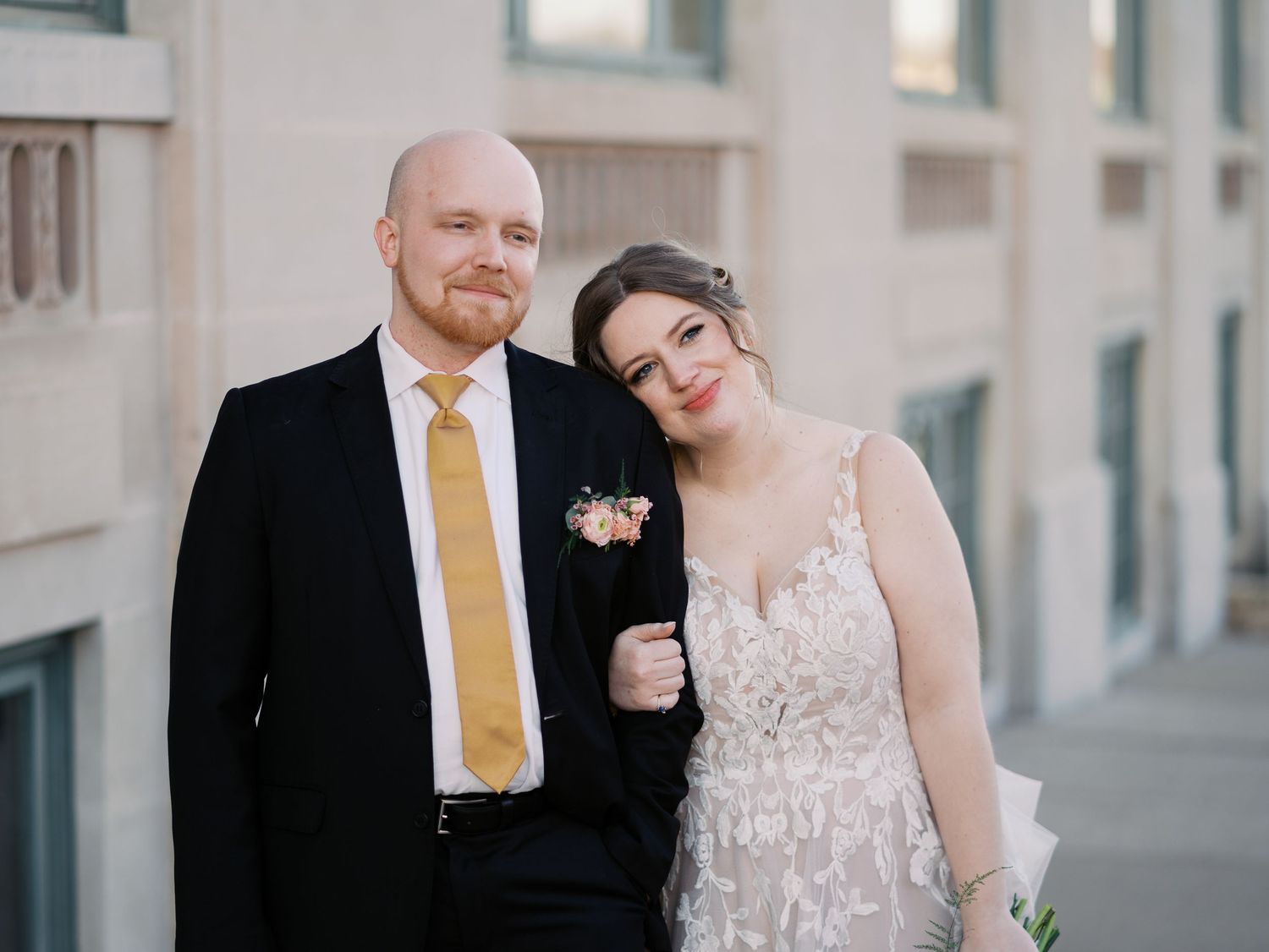 A couple poses together at their wedding, with the bride in a lace dress and groom wearing a yellow tie.