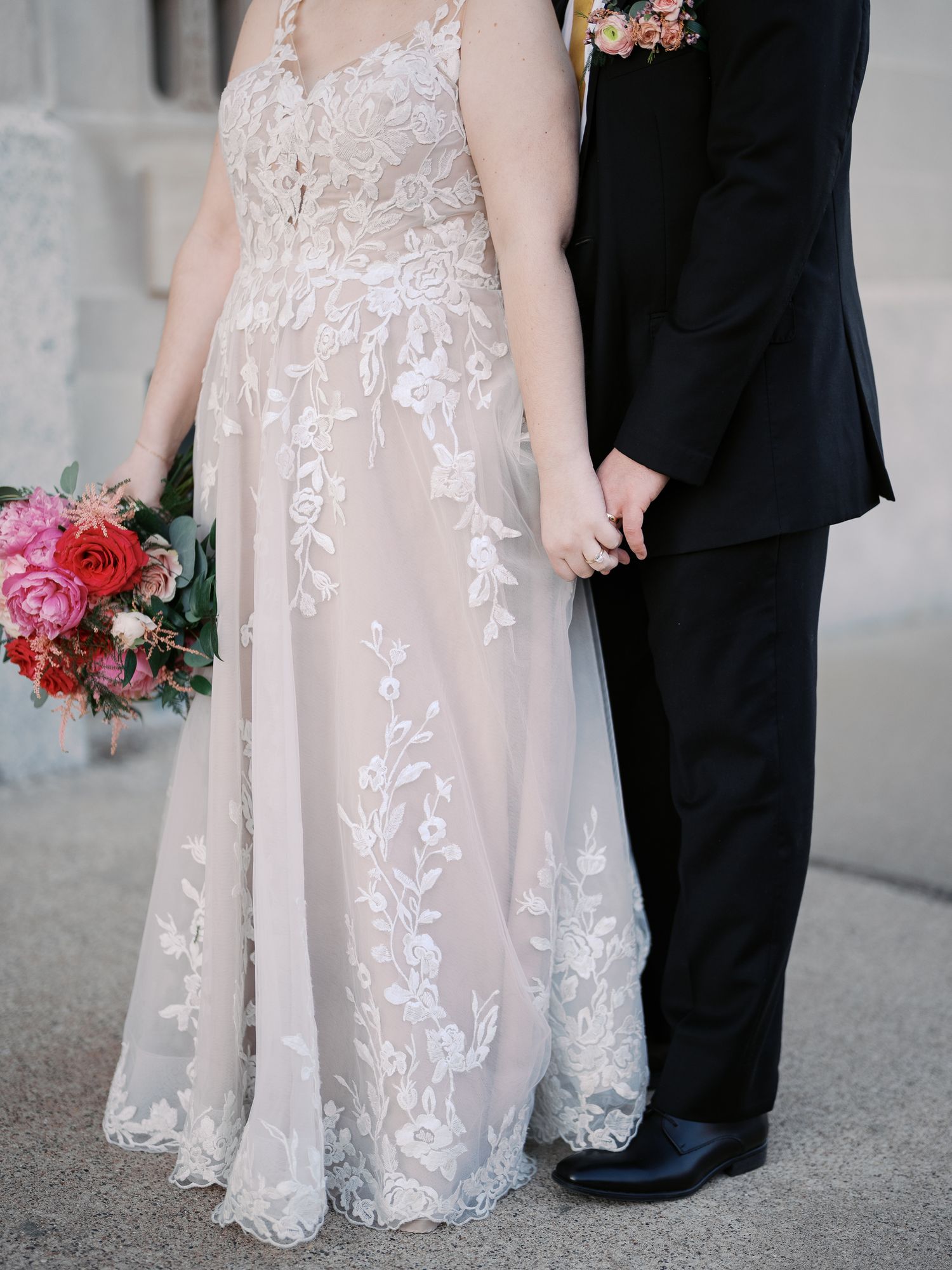 A detailed close-up of a bride in a lace wedding gown holding a pink bouquet next to a groom in a black suit.