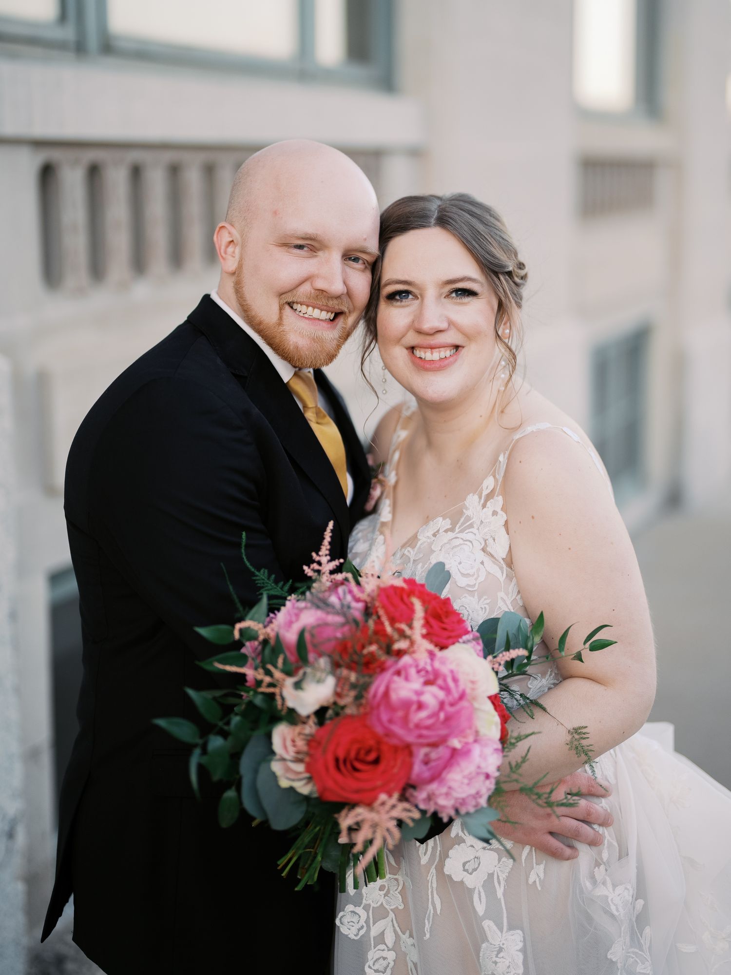 A newlywed couple in formal attire pose together with a vibrant pink and red floral bouquet at their wedding.