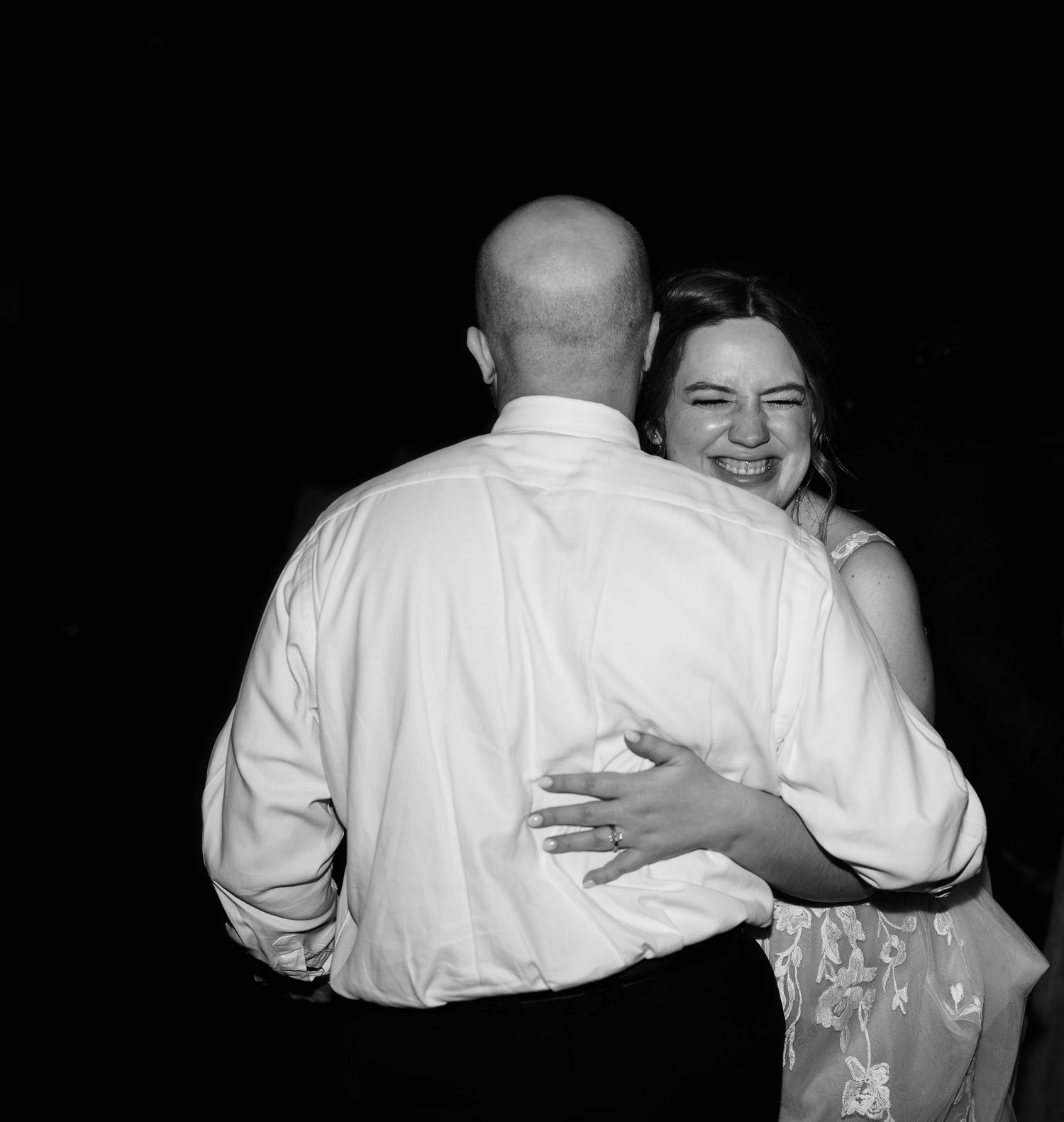 A black and white photo of a couple embracing while slow dancing at what appears to be a wedding reception.