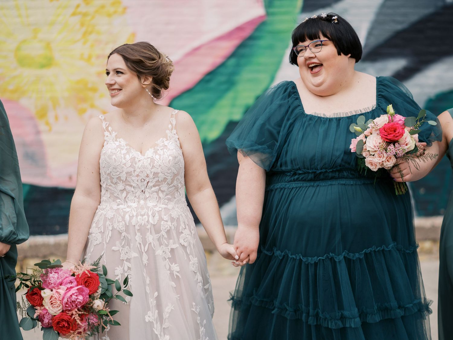 Two happy people in wedding attire holding pink bouquets stand together holding hands in front of a colorful mural.