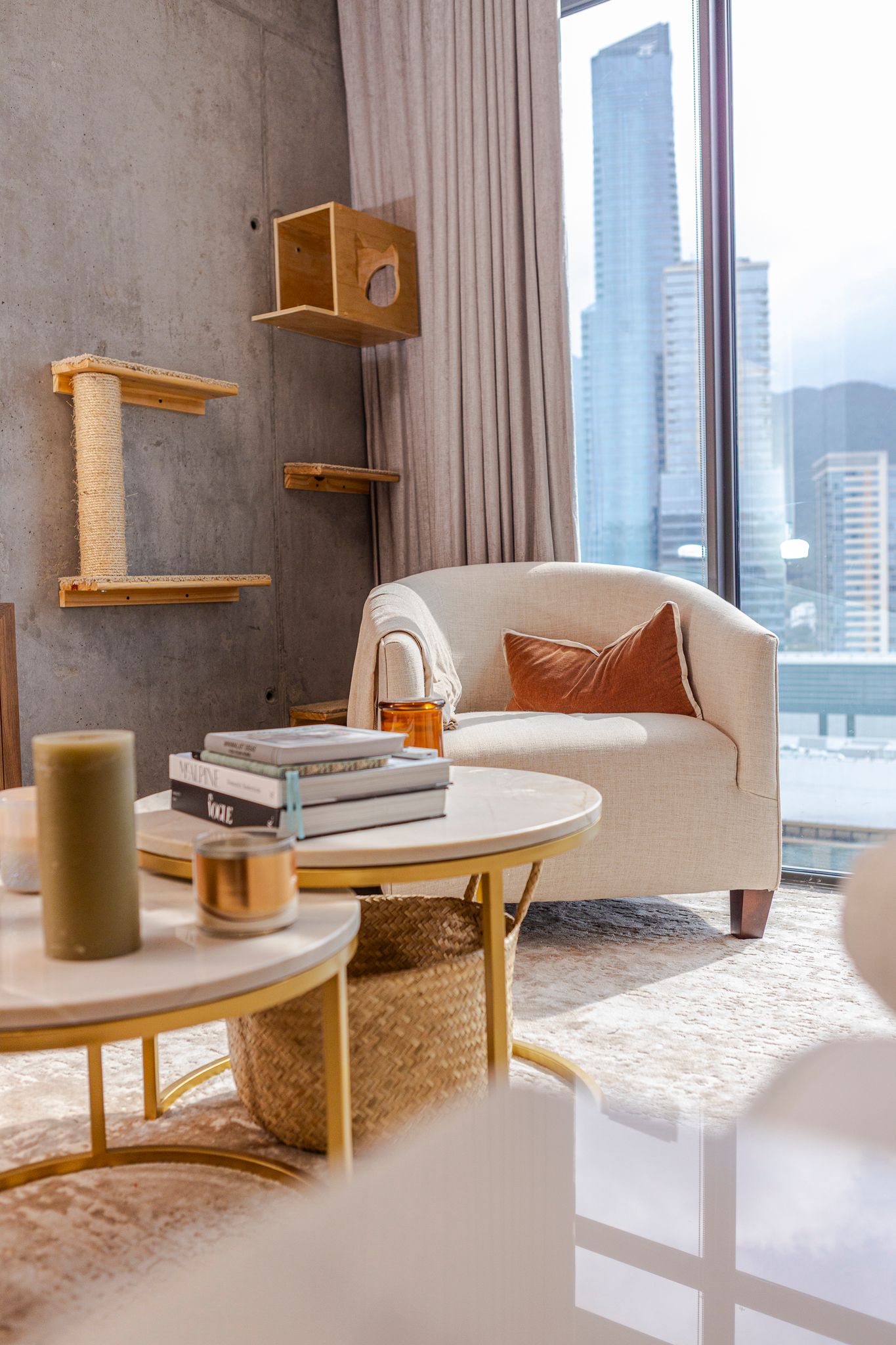 Modern living room with cream armchair, wooden shelves, and city view through window displaying urban high-rise buildings.