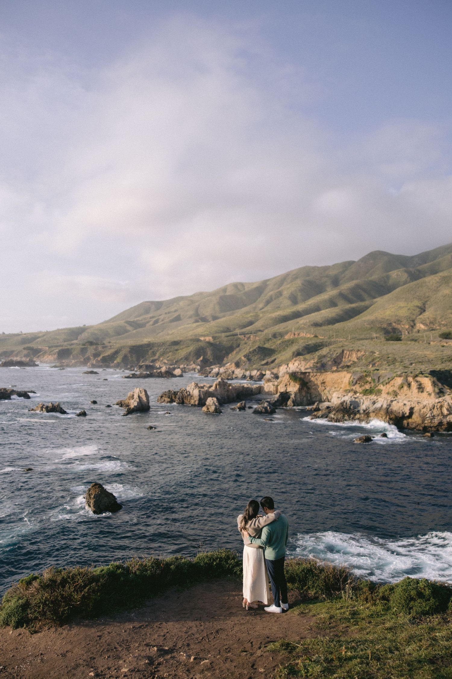 A couple embraces on a cliffside overlooking the dramatic Big Sur coastline with rocky shores and rolling hills in California.